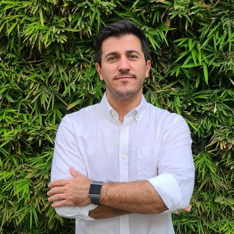 A man with dark hair and a trimmed beard wearing a white button-up shirt and a black watch, standing in front of lush green foliage, with his arms crossed.