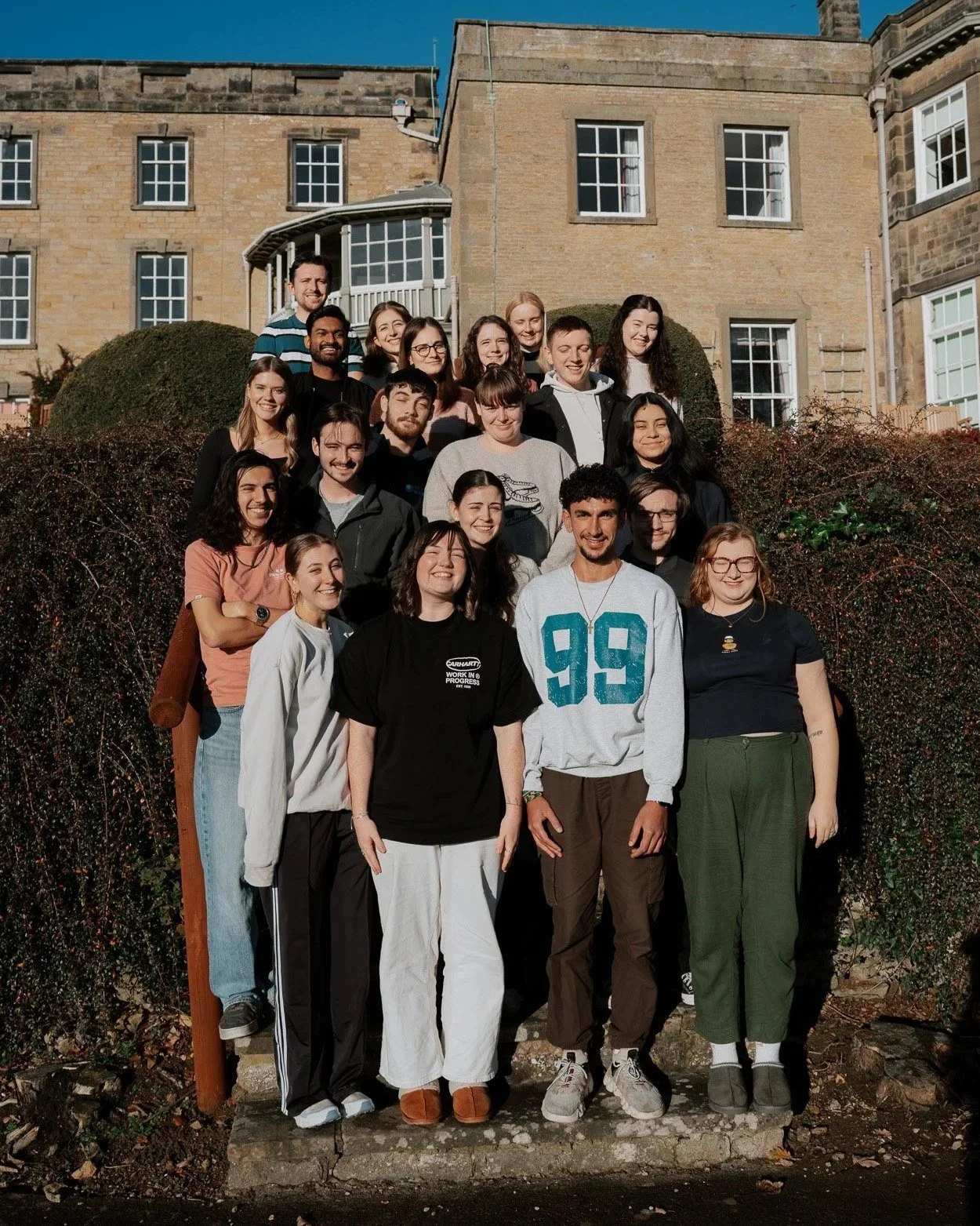 A group of 21 young adults gathered outdoors on steps, smiling, in front of a large stone building with multiple windows under a blue sky.