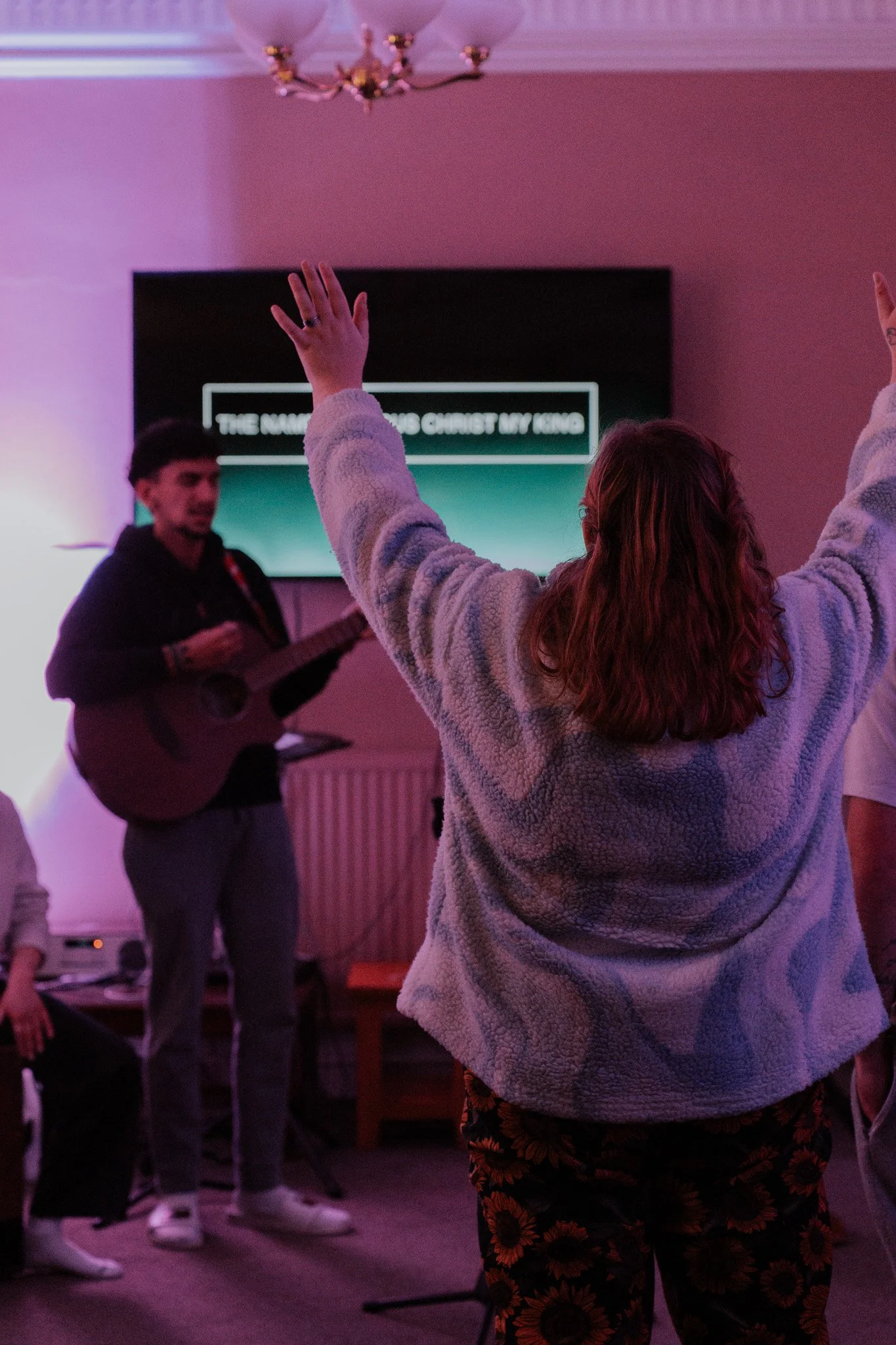 A woman with red hair, wearing a fuzzy gray jacket and floral pants, raises her arms during a worship session as a man in a black hoodie plays guitar in a dimly lit room with purple and pink lighting and a screen behind them displaying the words 'The Name of Jesus Christ My King'.