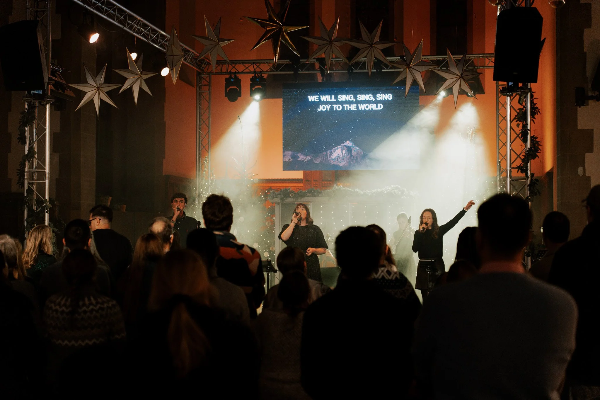 People attending a live Christmas concert or worship event with singers on stage, decorated with Christmas lights and stars, and song lyrics displayed on a large screen.