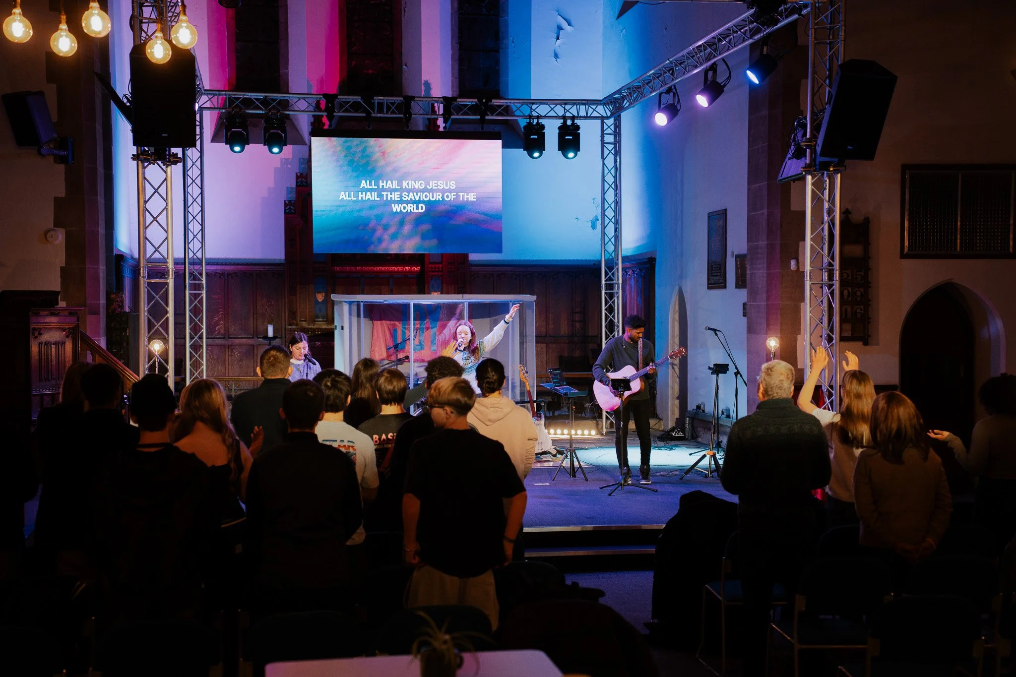 A worship band performs on stage in a church with a choir singing, while the congregation stands and raises hands during a service. The stage has a large screen displaying lyrics, and colorful lighting enhances the ambiance.
