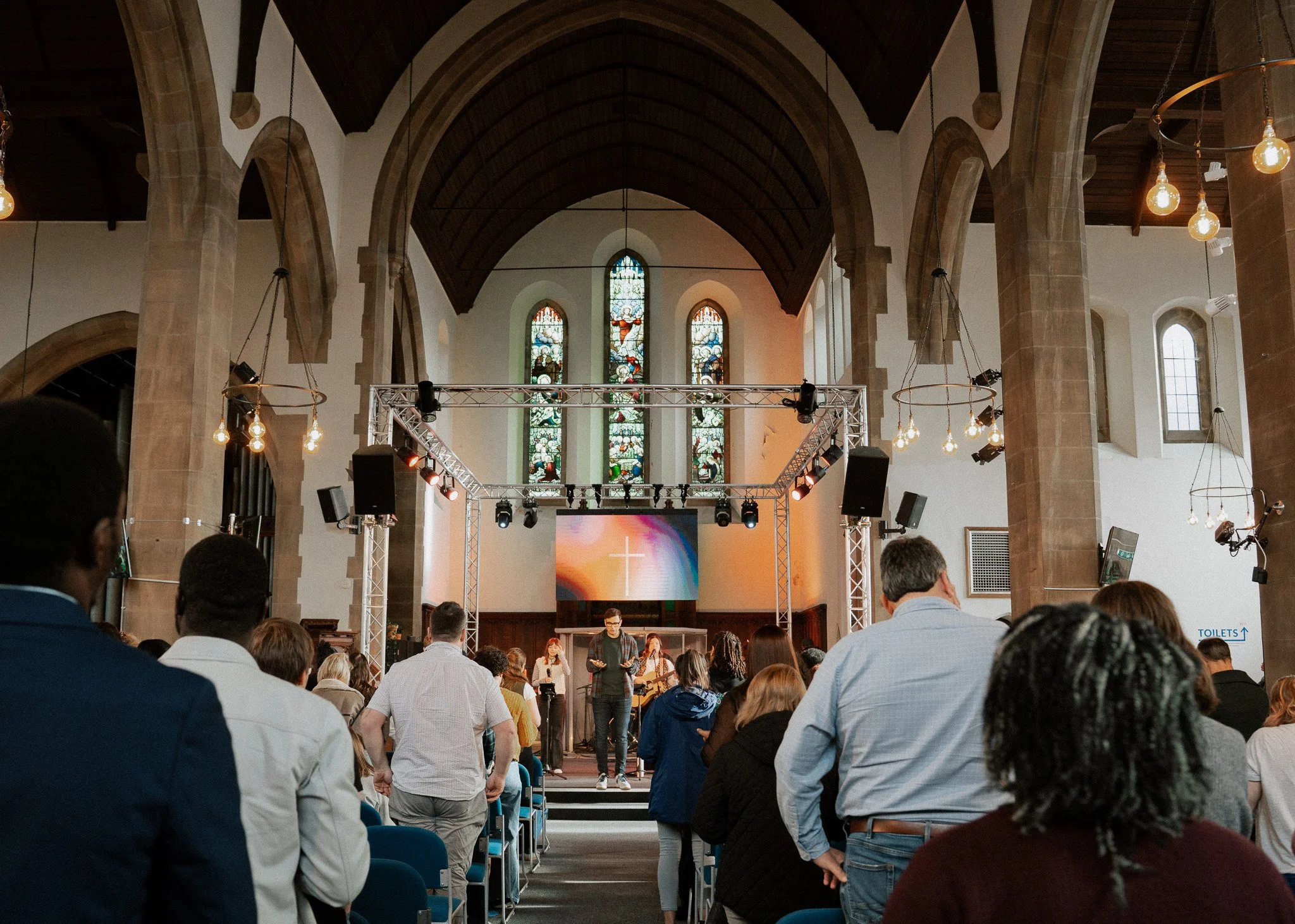 A congregation inside a church during a service or event, with a stage at the front featuring musicians and a large screen with a cross symbol, stained glass windows behind the stage, and people standing and seated.