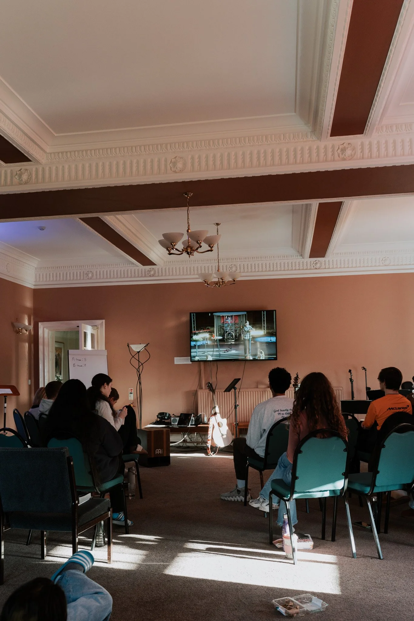 Group of people watching a presentation in a room with ornate ceiling, chandelier, and large TV screen.
