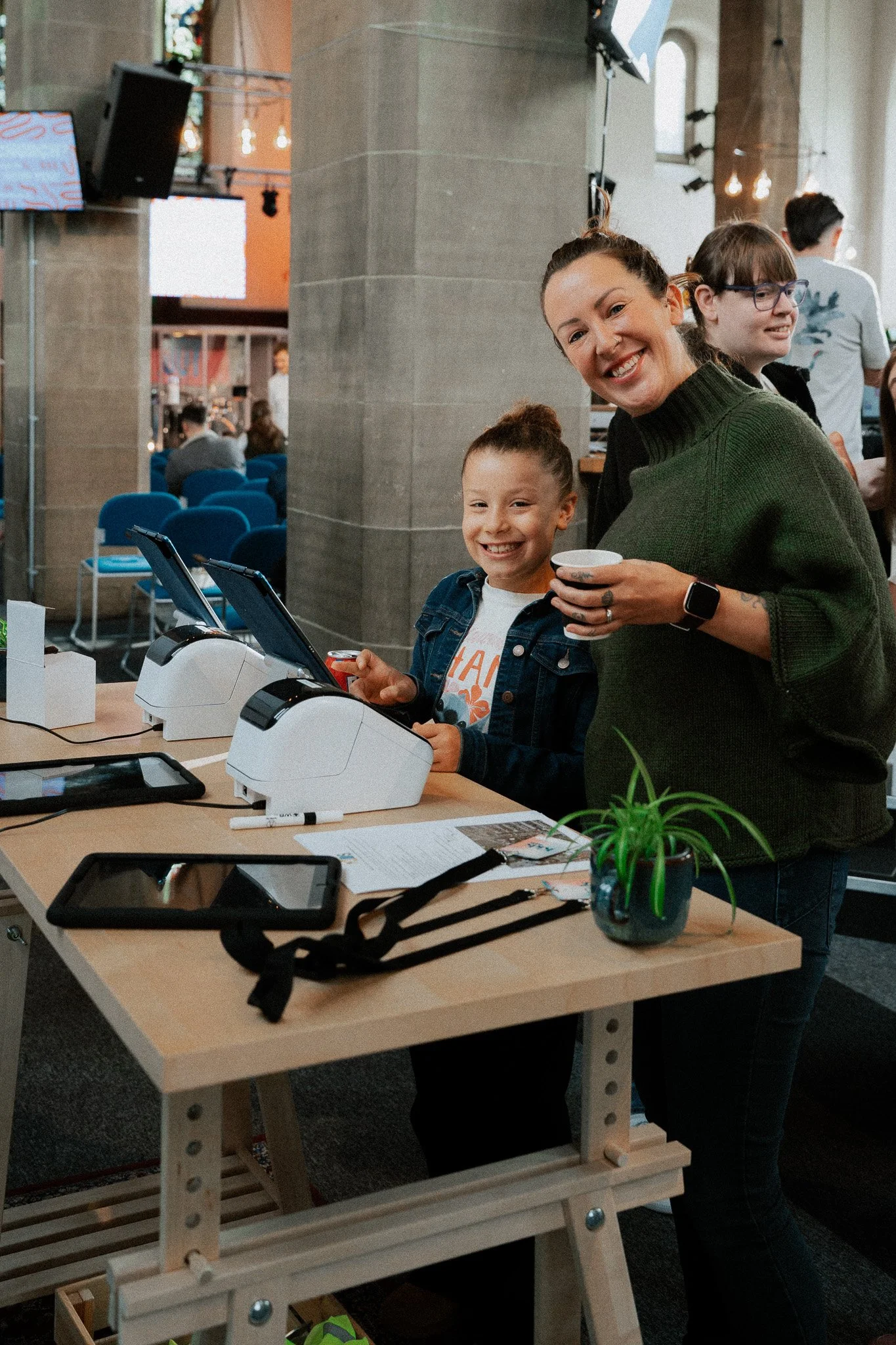 A woman and a girl smiling at a table with two small barcode scanners, tablets, and a potted plant in a busy indoor setting.