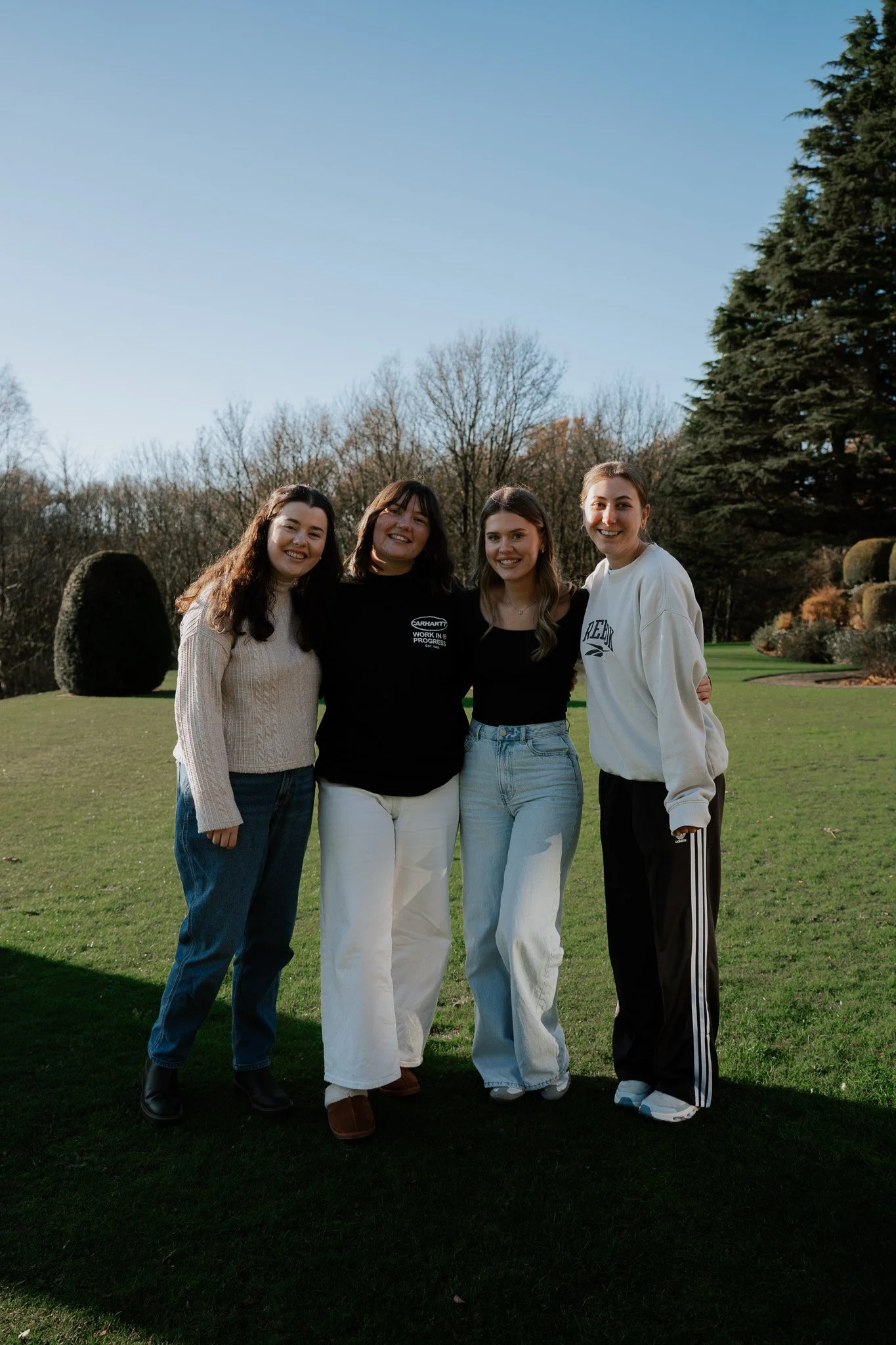 Three young women standing together outdoors on a grassy field, smiling at the camera with trees in the background.