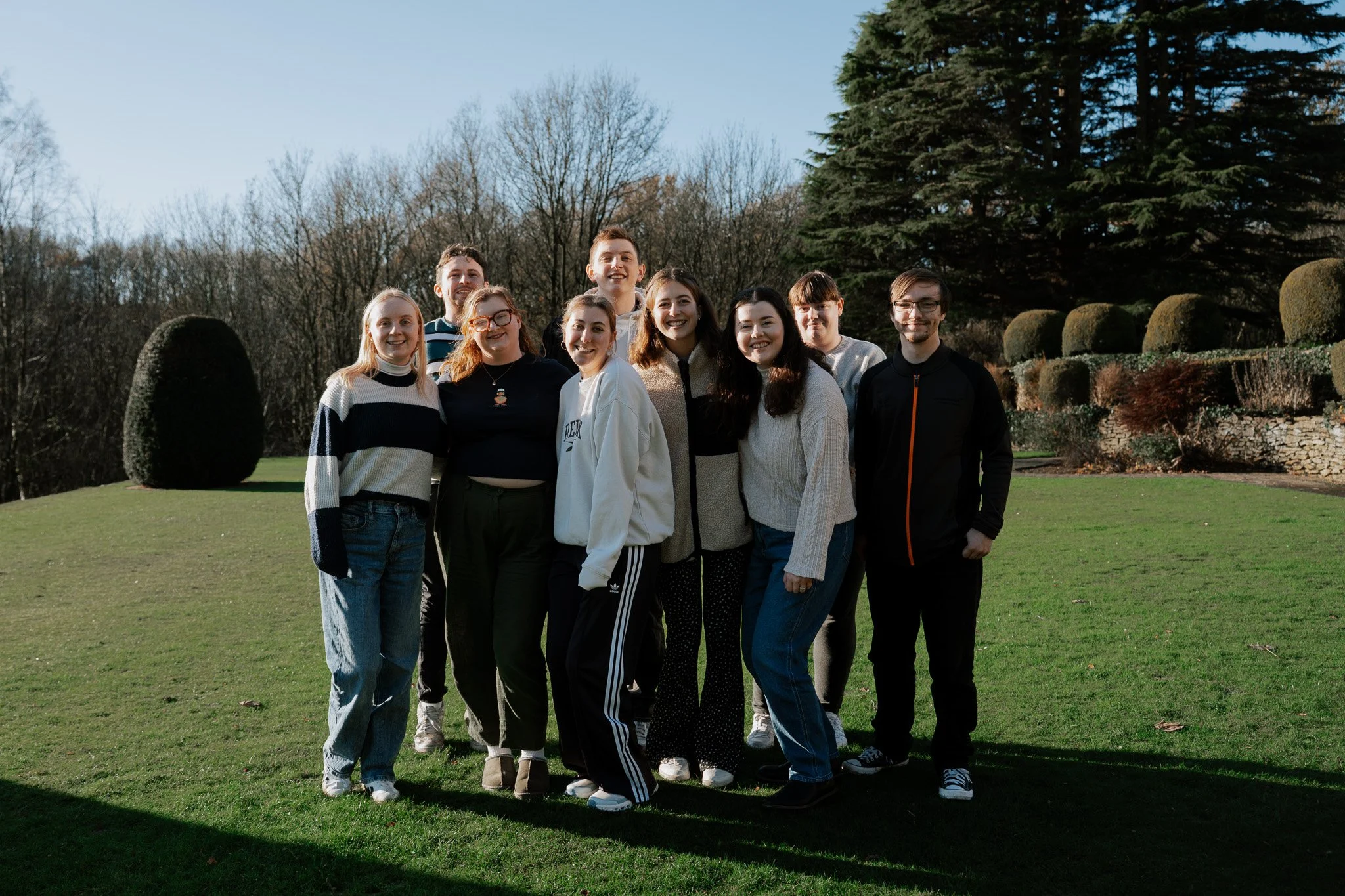 A group of ten young people standing outdoors on a grassy area, smiling for a photo, with trees and bushes in the background during daytime.