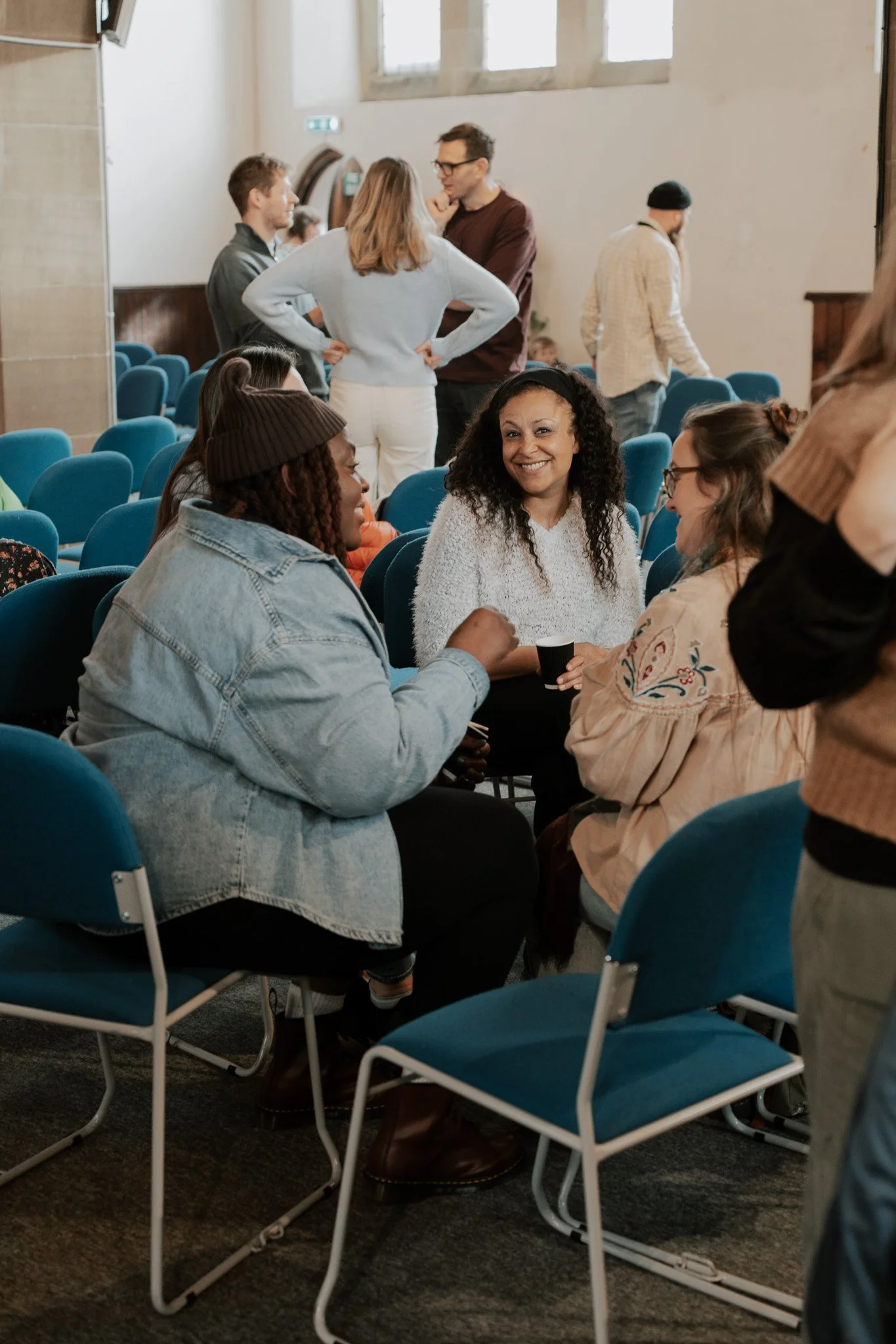 Group of people in a room with blue chairs, engaging in conversation and smiling, with some standing and some sitting.