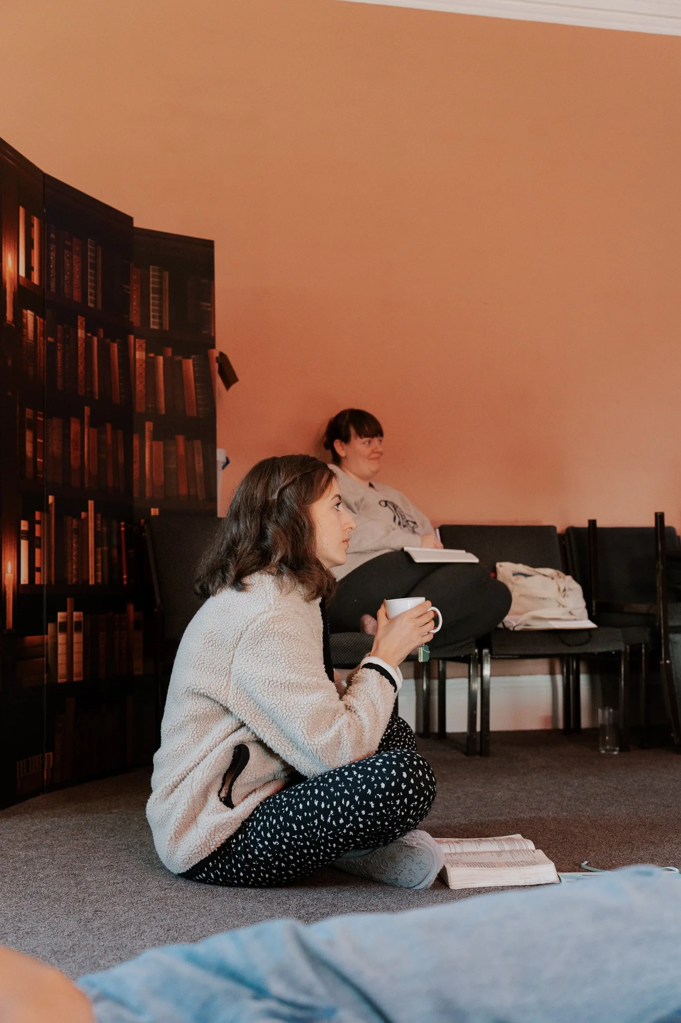 A woman sitting cross-legged on the floor, holding a mug, with a book open in front of her. In the background, another woman sits on a chair, listening and taking notes. The setting appears to be a cozy indoor space with a bookshelf wallpaper design on the wall.