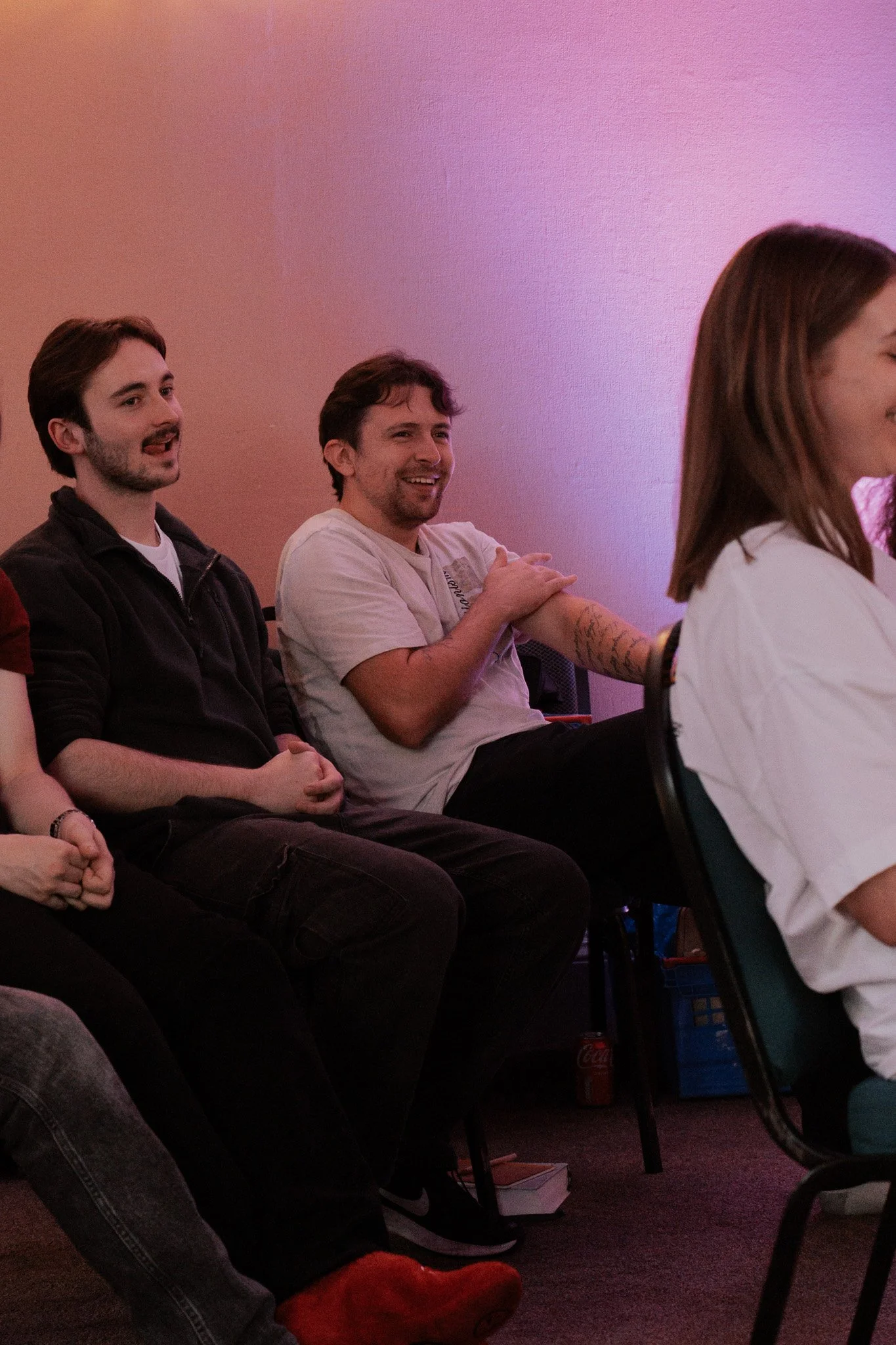 People sitting and looking towards the front, smiling and enjoying a presentation or performance in a room with pink lighting.