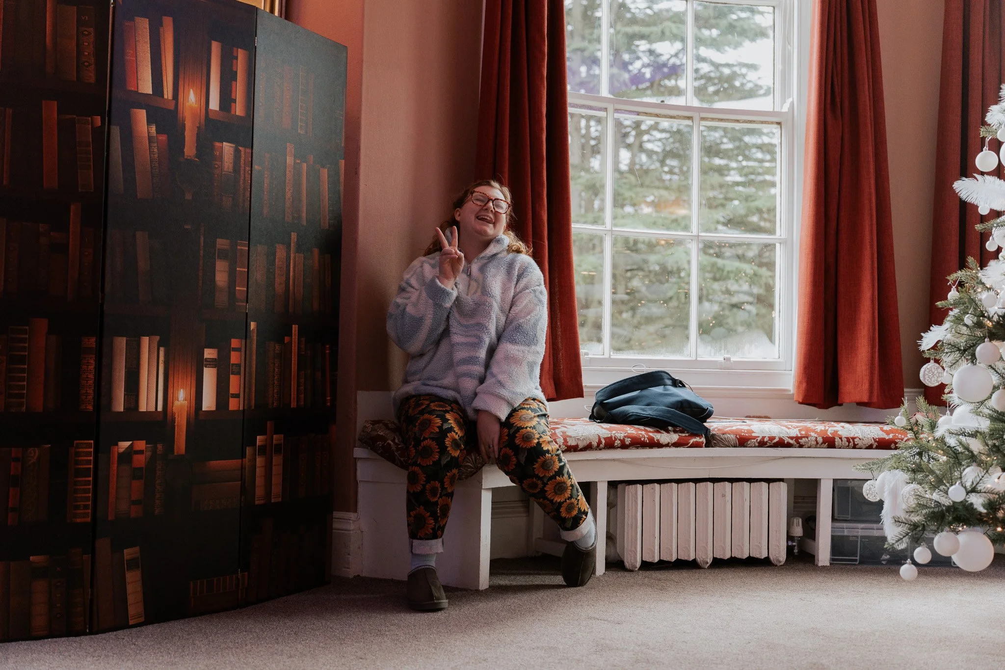 A girl sitting on a window seat in a cozy room, smiling and making a peace sign with her fingers.