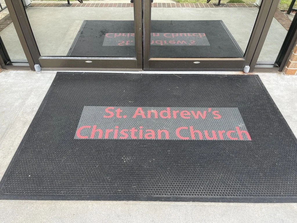 Black doormat with red letters reading 'St. Andrew's Christian Church' in front of glass doors.