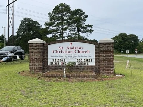 Sign for St. Andrews Christian Church on a brick and white signboard with welcome message and service times, surrounded by grass and trees.