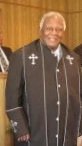 Official dressed in black clerical attire with cross pins, smiling in front of a wooden background.