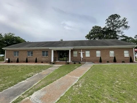 A single-story brick church building with a cross on the front, surrounded by a well-kept lawn and a sidewalk leading to the entrance, under a cloudy sky.