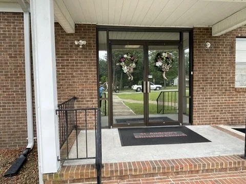 Glass door entrance decorated with two floral wreaths, brick building exterior, black railing, and a welcome mat.