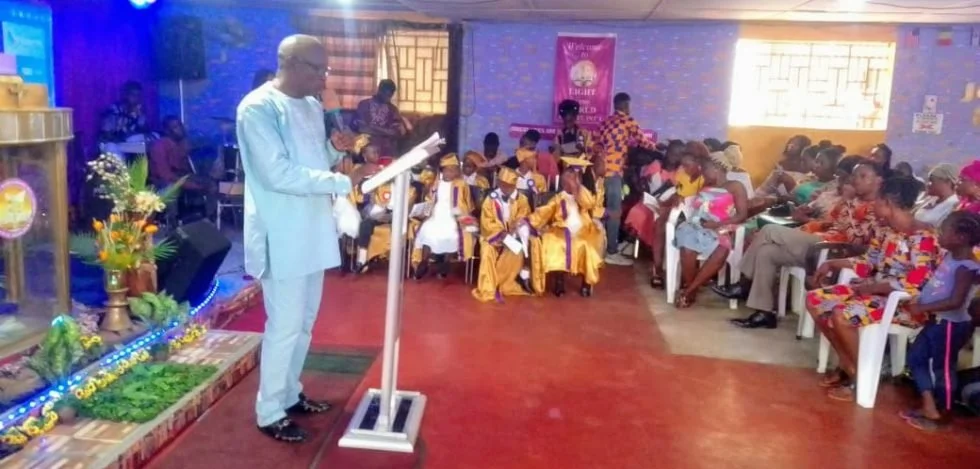 Man dressed in white delivering a speech at a podium during a ceremony, with children in graduation gowns and caps seated and clapping in an indoor hall decorated with flowers and colorful banners.