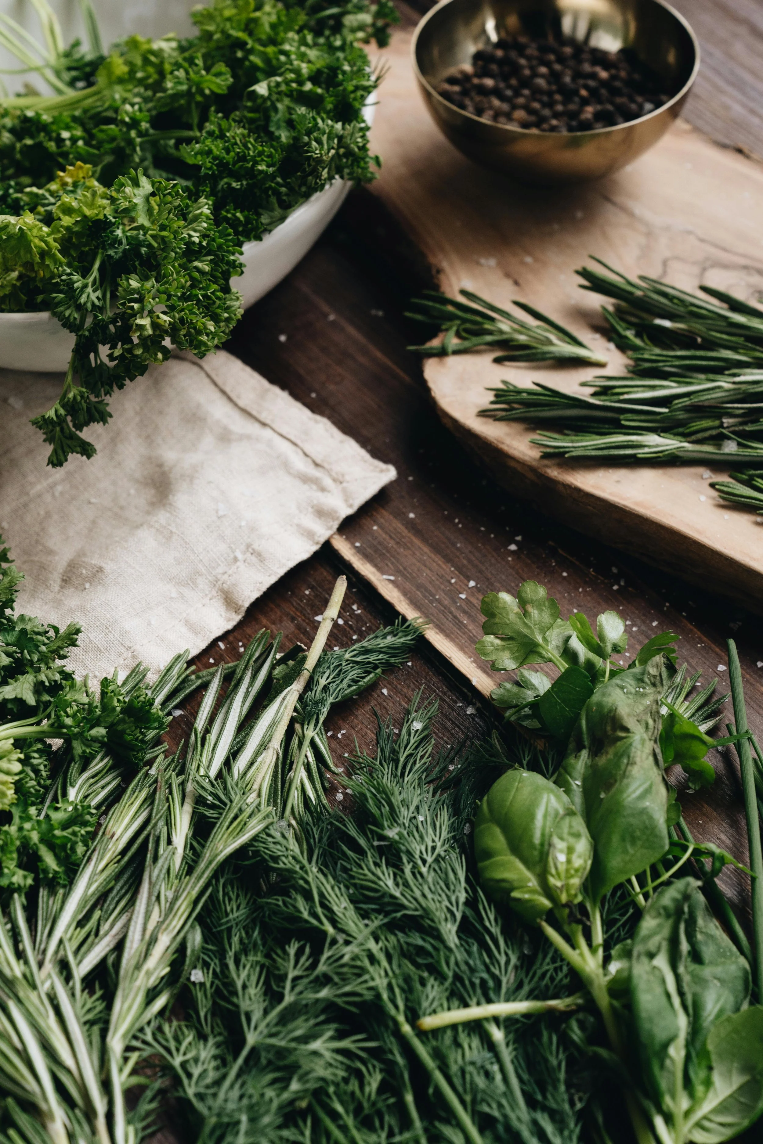 Various fresh herbs including parsley, rosemary, dill, and basil on a wooden table with a bowl of black peppercorns in the background.