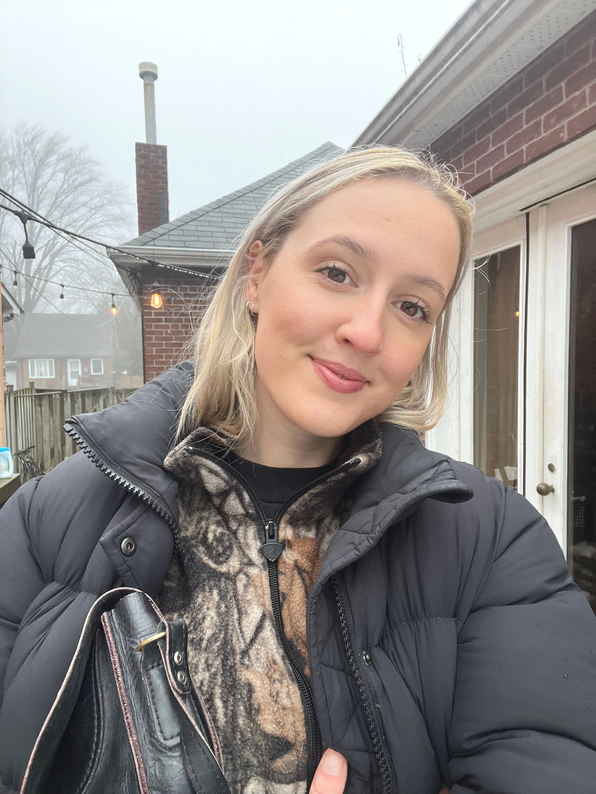A young woman with blonde hair taking a selfie outside on a cloudy day. She is wearing a black puffer jacket, a patterned fleece, and is holding a black handbag. There is a brick house with a sliding glass door behind her, and in the background, an outdoor patio with string lights and a wooden fence.