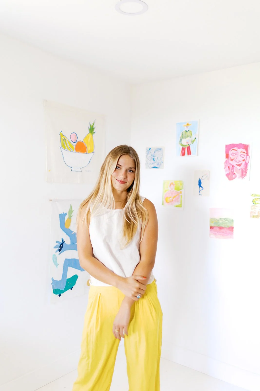 Young woman with long blonde hair wearing a white sleeveless top and yellow pants, standing in an art gallery with colorful drawings on white walls.