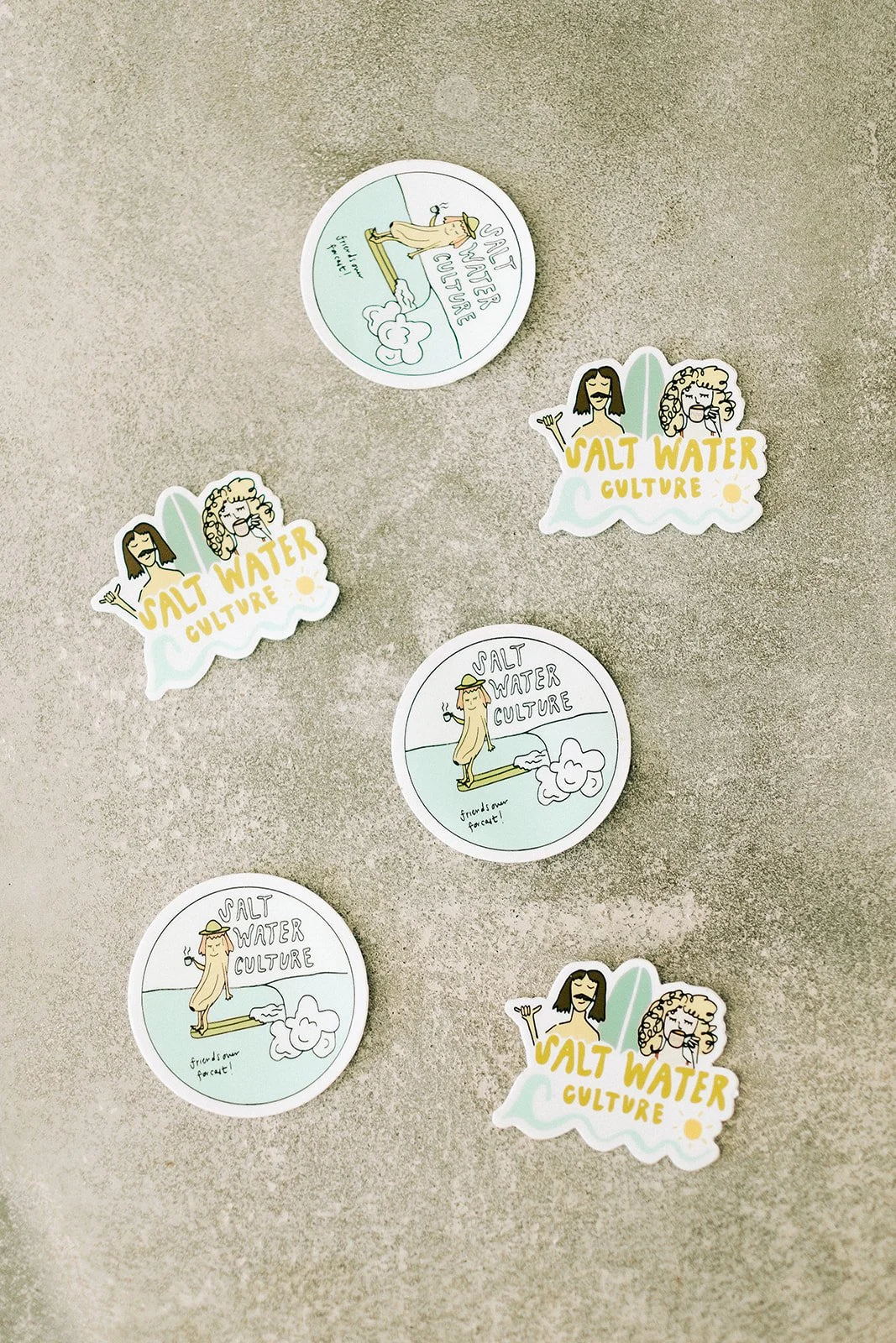Four stickers on a textured surface, with three stickers reading "Salt Water Culture" showing cartoon women with surfboards and waves, and one sticker with a cartoon woman in a hat with a wave and the text "Friend or foe!".