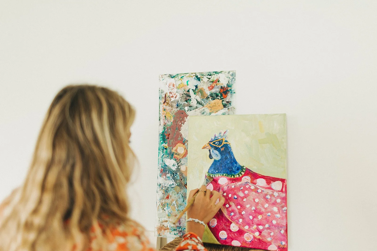 A woman with long blonde hair painting on a canvas that depicts a colorful peacock with a pink polka-dotted tail, in front of a white wall.