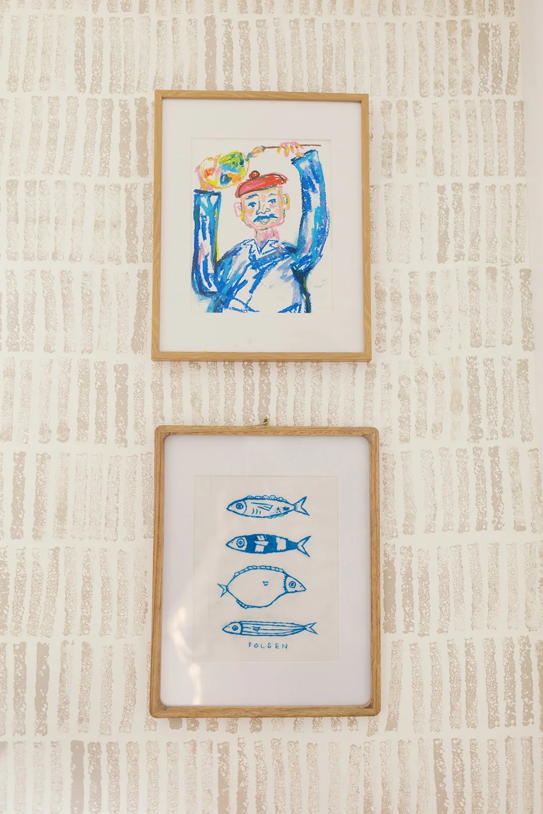 Two framed artworks on a beige textured wall; the top artwork is a colorful, childlike painting of a man with a mustache, red hat, and blue clothing holding a yellow object; the bottom artwork is a blue line drawing of various fish and the word "FOLS