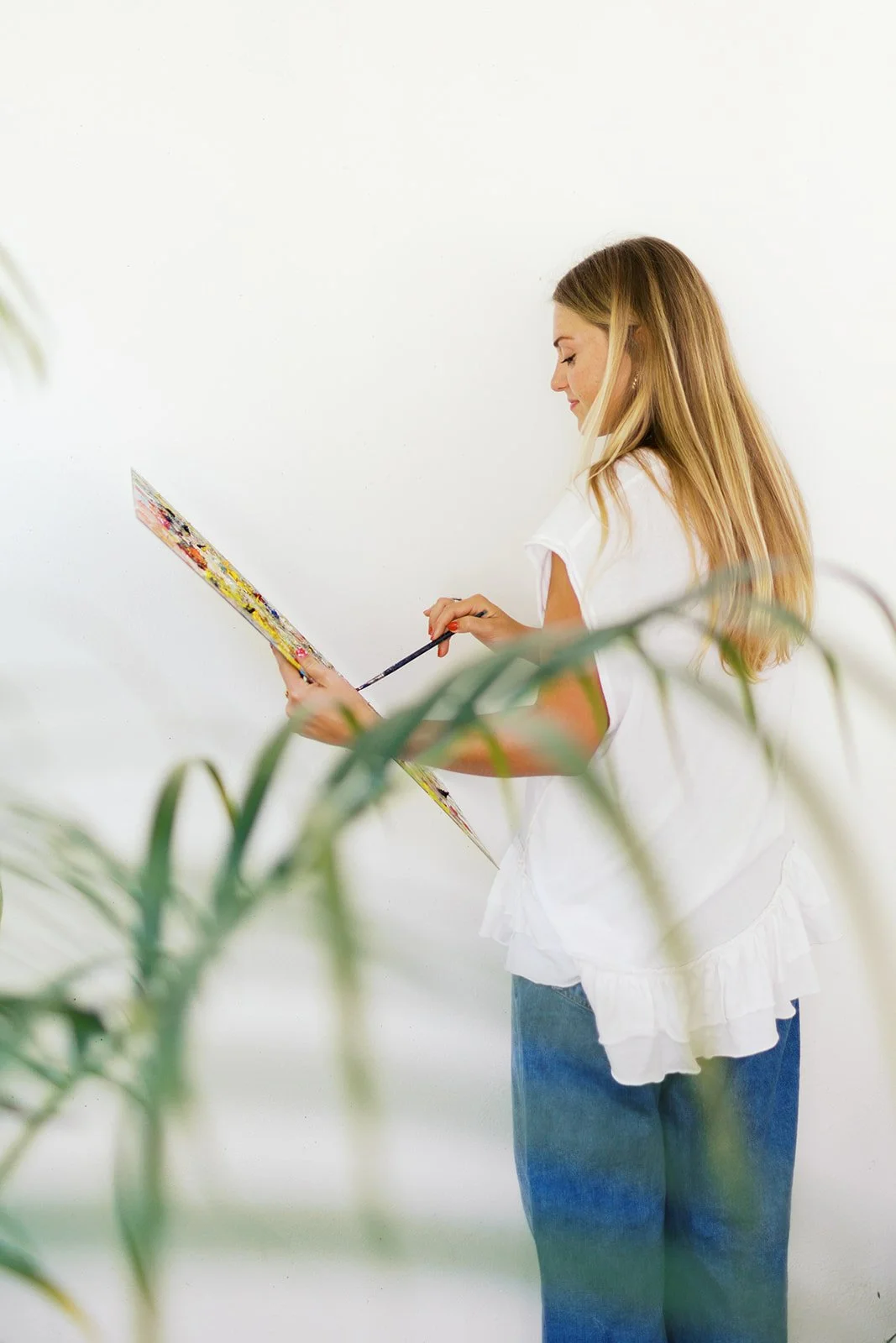 A woman with long blonde hair, wearing a white ruffled blouse and blue jeans, is holding a paint palette and brush, standing against a white background, partially obscured by green plants in the foreground.