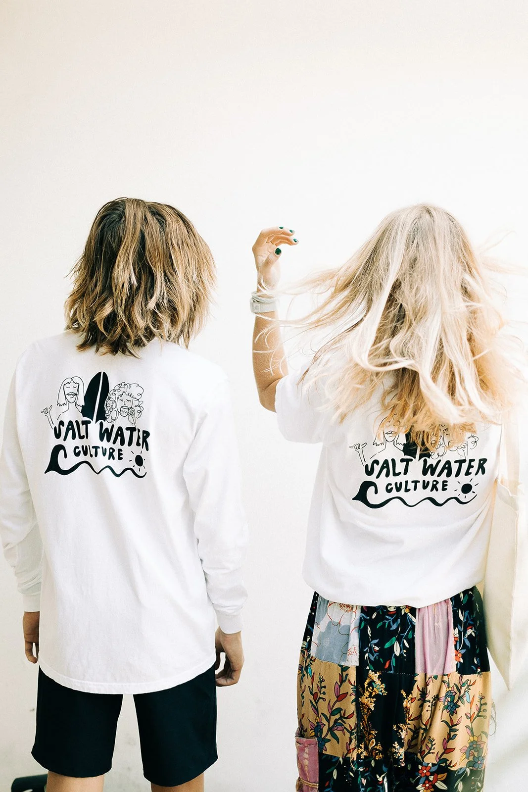 Two women with blonde hair, one with short hair and the other with long, wavy hair, wearing white t-shirts with a logo that says "Salt Water Culture" and an illustration of two women and waves, standing against a plain white wall.