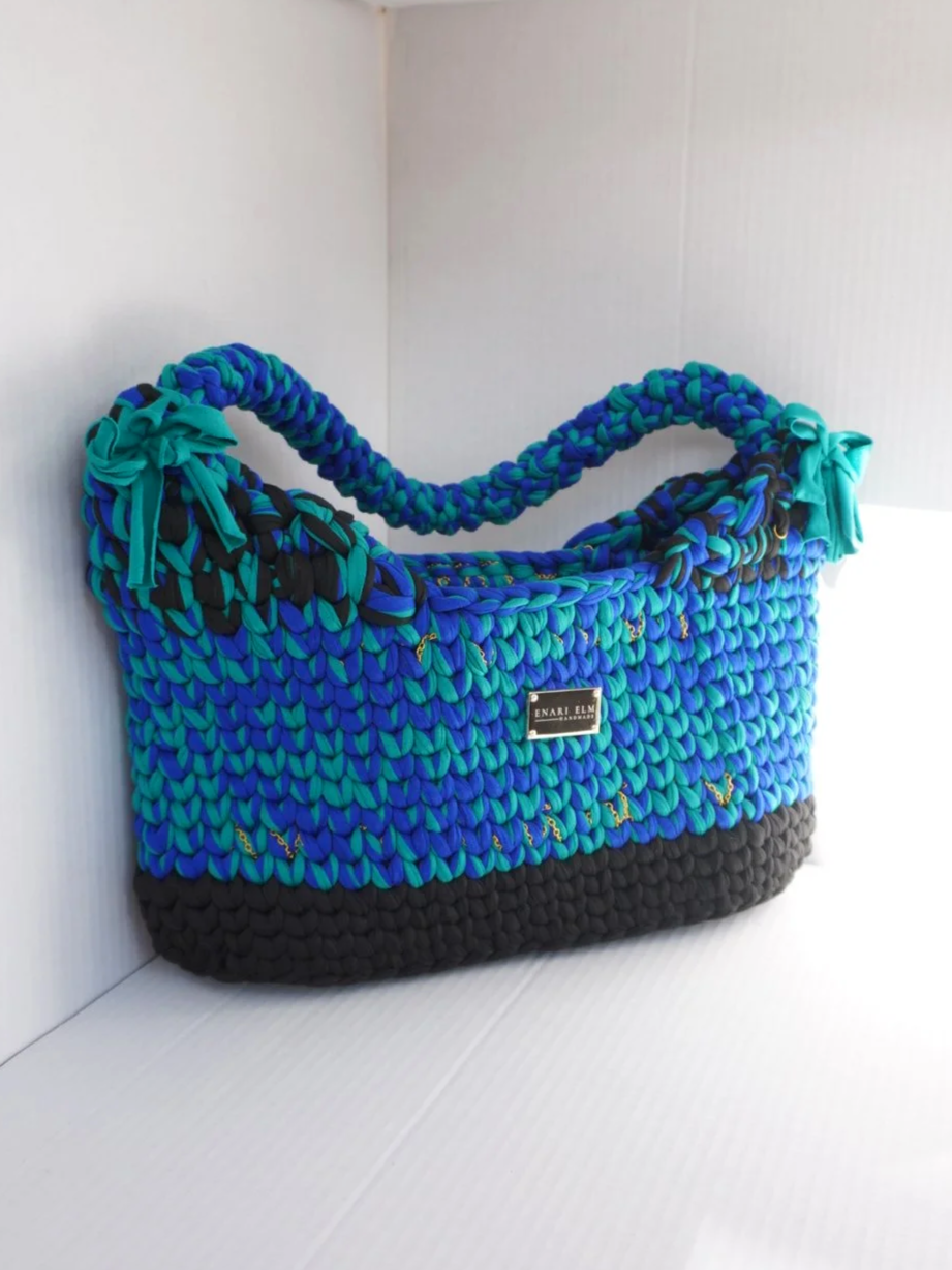 Hand-crocheted shoulder bag in teal, blue, and charcoal with braided strap and textured stitching on a white background