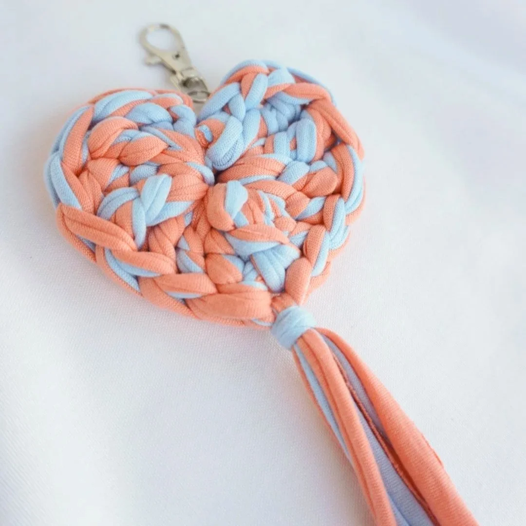 Alt Text Peach and light blue crochet heart keychain with a soft fabric tassel on a silver lobster clasp, photographed on a white fabric background.