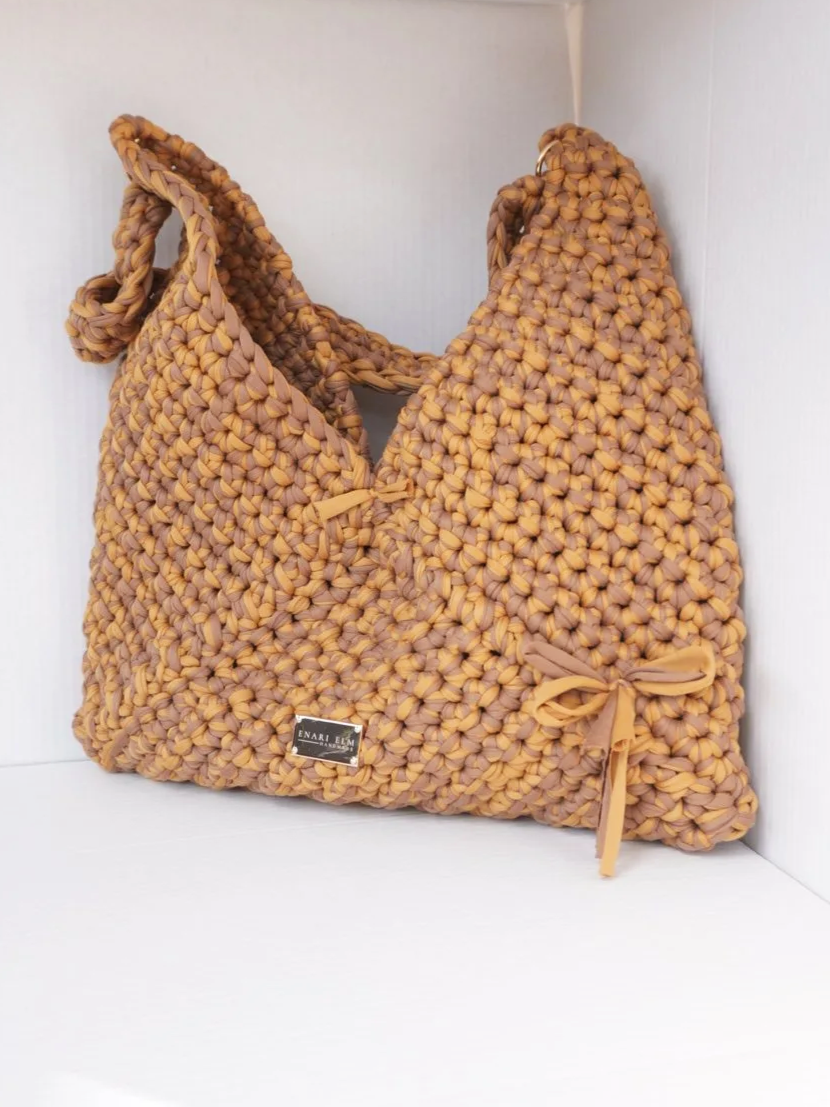 Hand-crocheted tan shoulder bag with soft textured stitching and tied bow detail, measuring 42 cm (16.5 in) long, 3 cm (1.2 in) wide, and 43 cm (16.9 in) tall