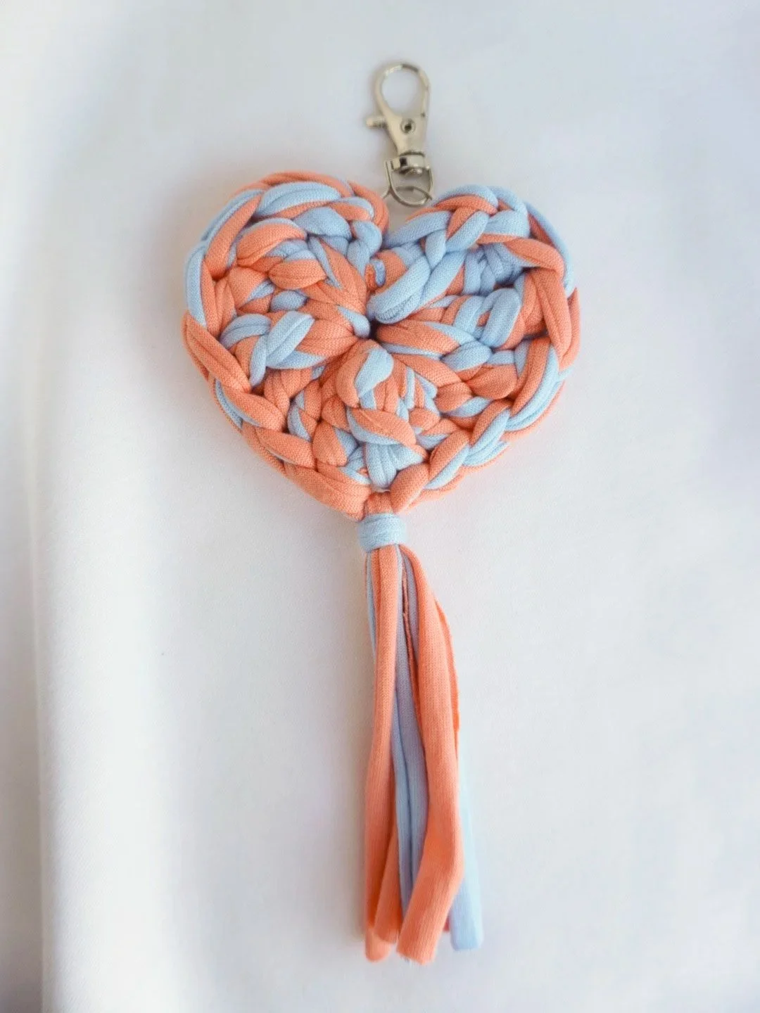 Alt Text Peach and light blue crochet heart keychain with a soft fabric tassel on a silver lobster clasp, photographed on a white fabric background.