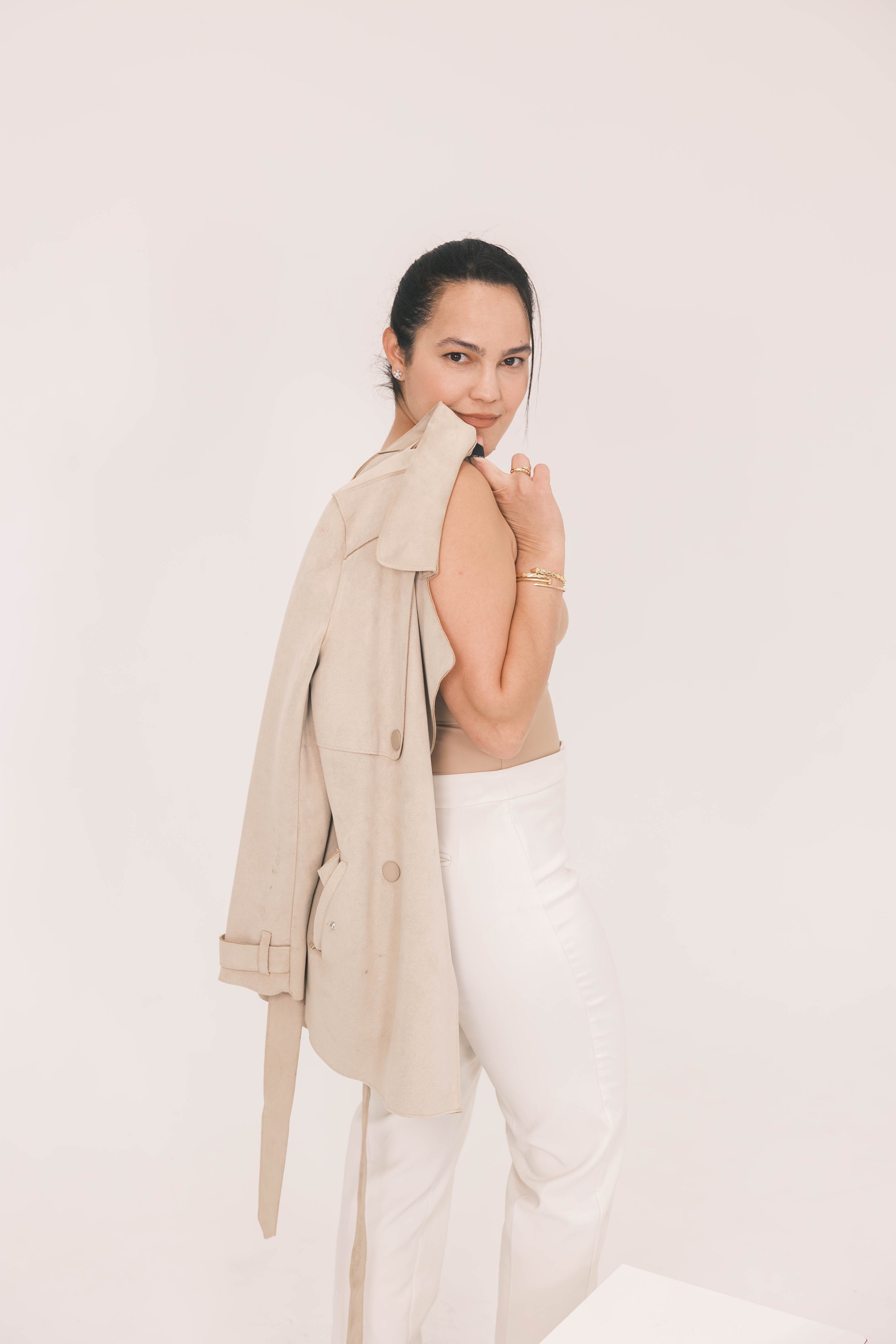 A woman with dark hair pulled back, wearing a beige sleeveless top and white pants, holding a beige trench coat over her shoulder, standing against a plain white background.