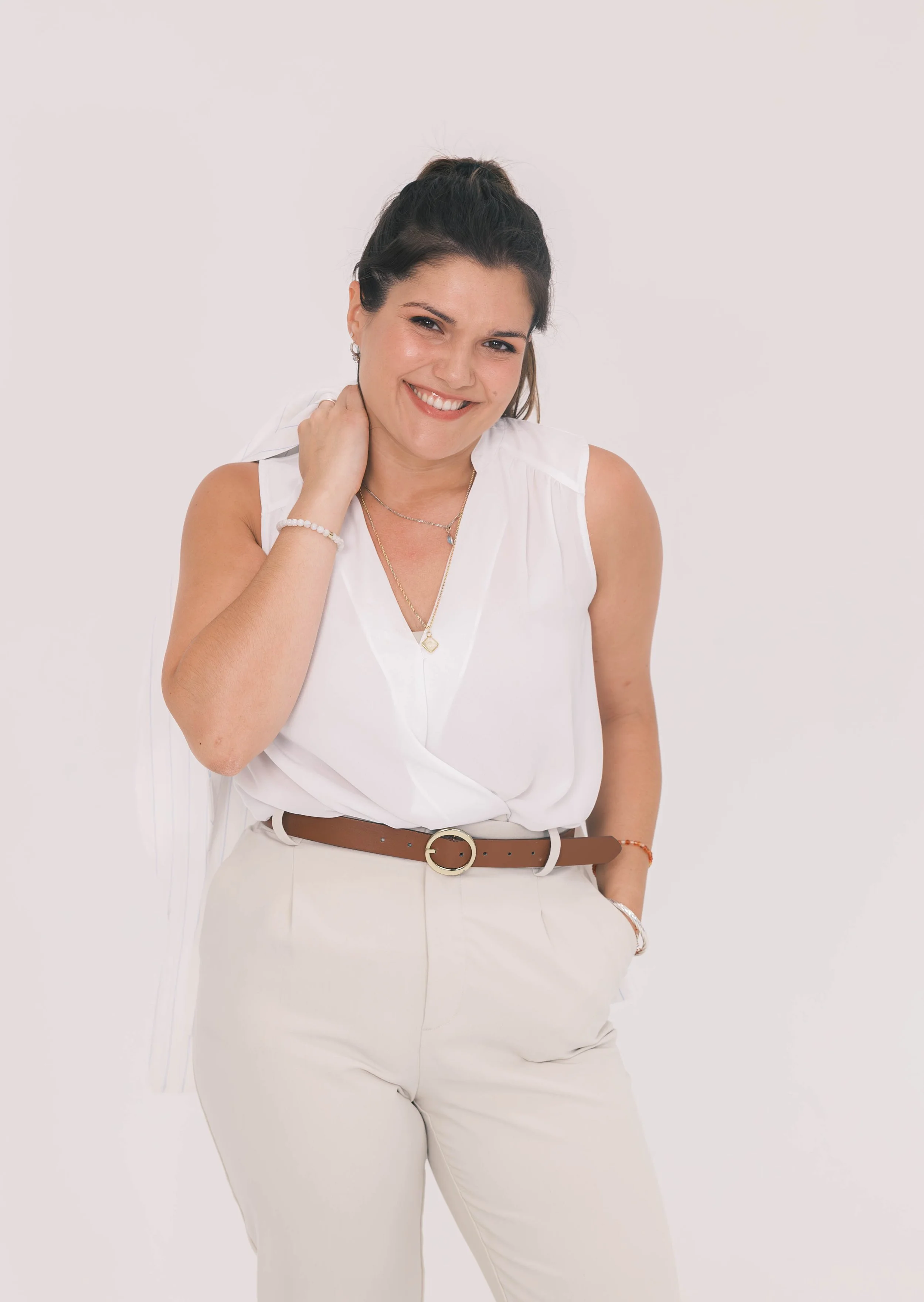 A woman with dark hair styled in a half-up, half-down look, wearing a white sleeveless blouse, cream-colored pants with a brown belt, and jewelry including a necklace, earrings, bracelets, and rings. She is smiling and has her right hand touching her neck. The background is plain white.