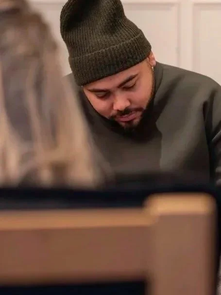 A young man wearing a dark beanie and gray sweatshirt looking down, possibly reading or writing, with a blurred person in the foreground.
