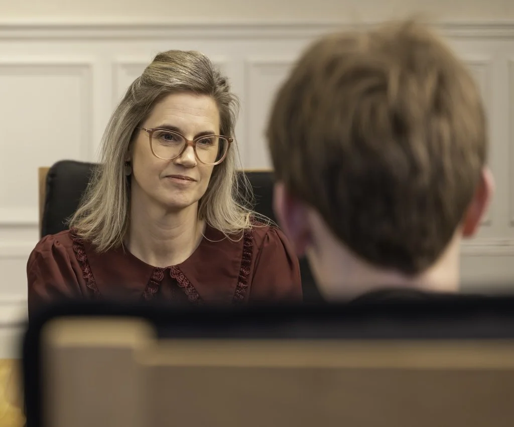 A woman with glasses and blonde hair wearing a red blouse, sitting in an office, interviewing a man with reddish hair.