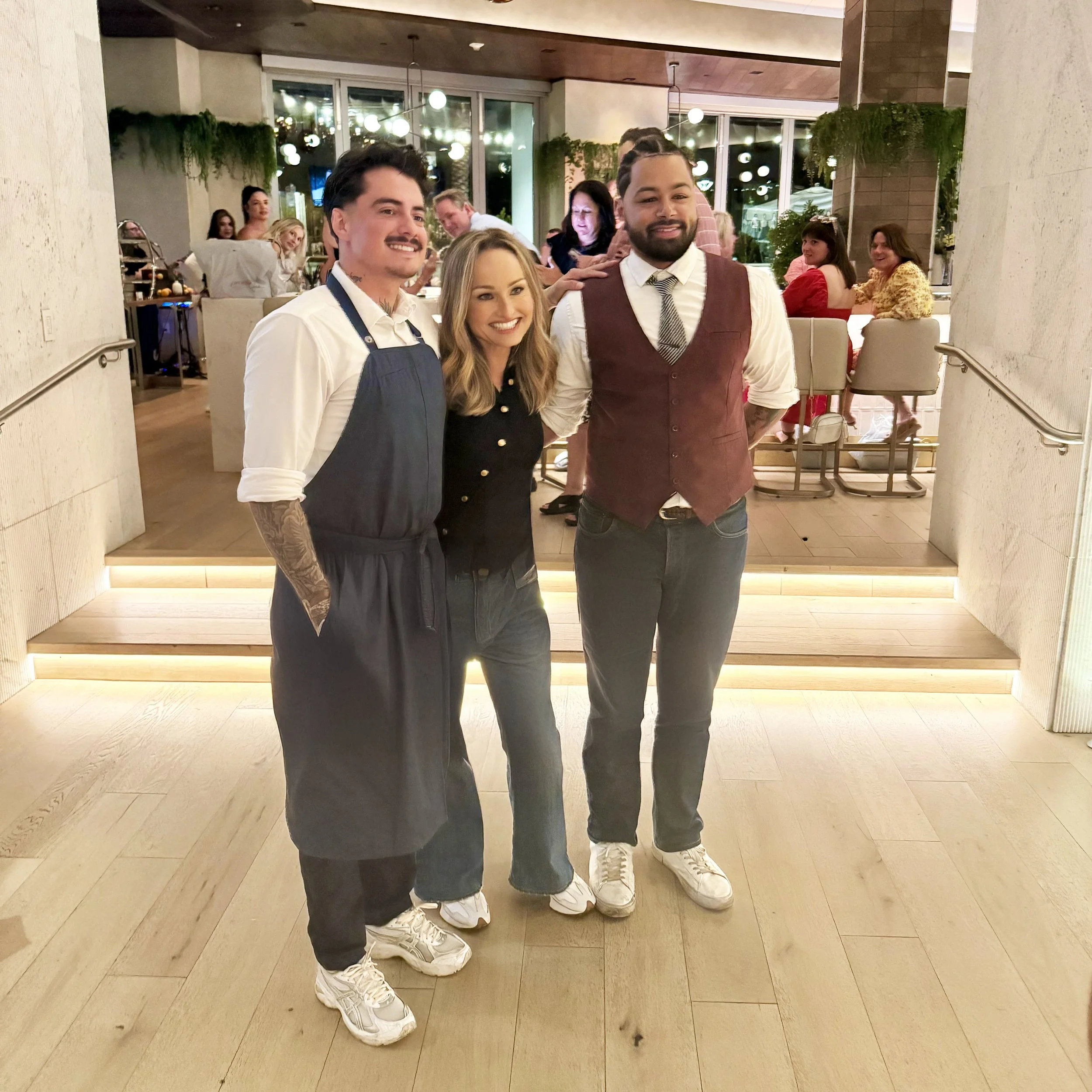 Three restaurant workers standing together, smiling, in a dining area with other patrons in the background at night.