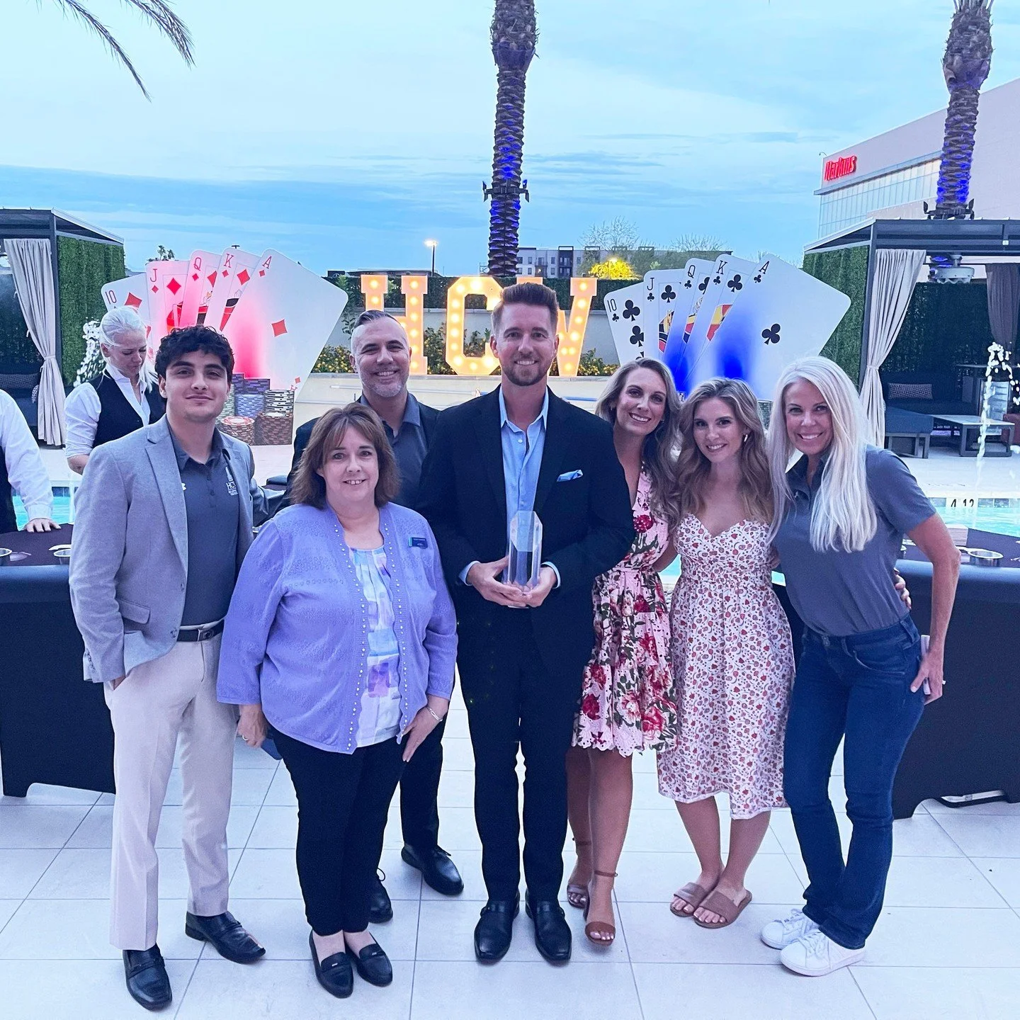 A group of seven people posing together outdoors at dusk, with a pool and decorative giant playing cards in the background, celebrating an event.