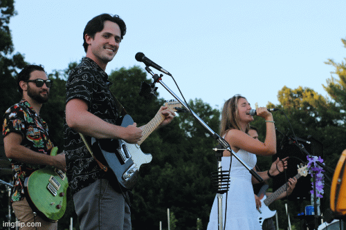 Band performing outdoors with three musicians playing guitars and singing, surrounded by trees.