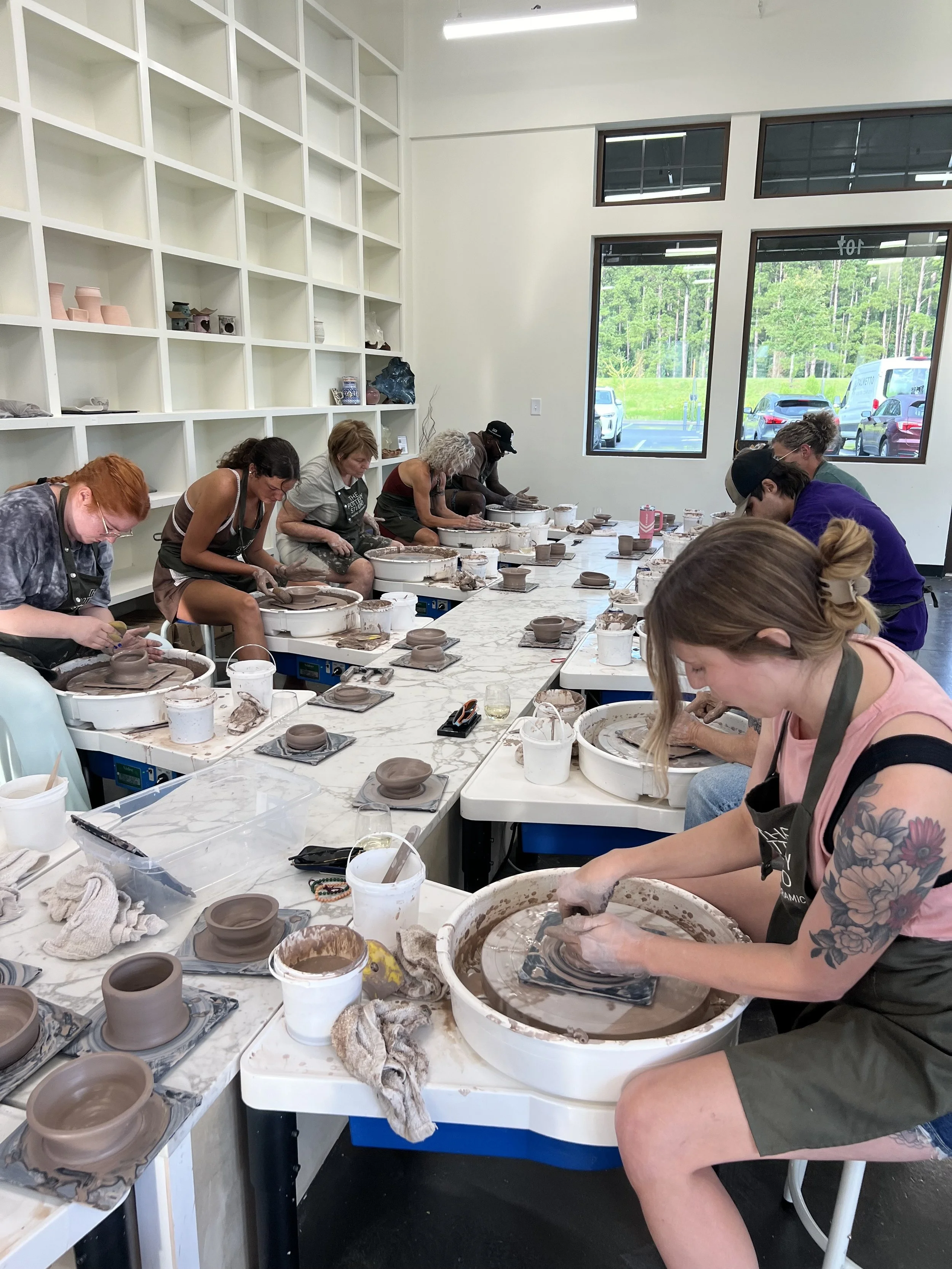 People participating in a pottery class, working on pottery wheels in a bright room with large windows and shelves with pottery pieces.