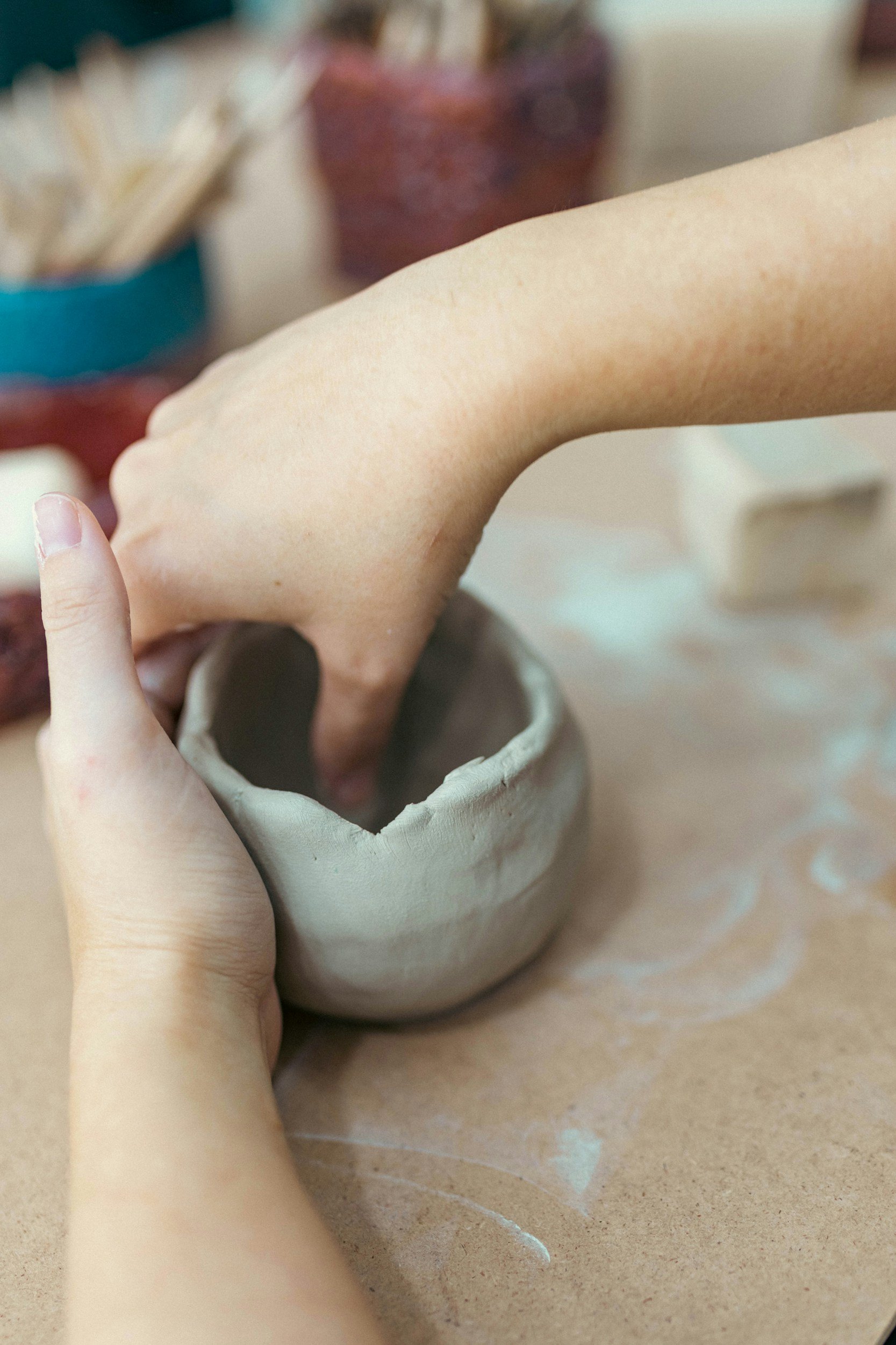 Two hands shaping a small clay pot on a work surface, with pottery tools and supplies in the background.
