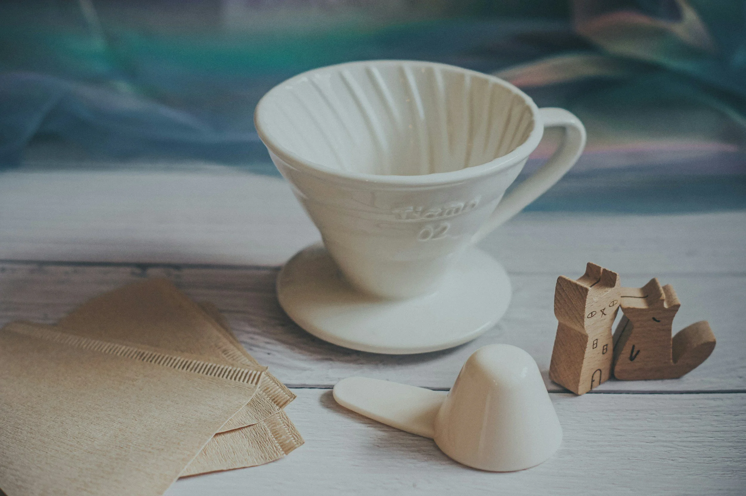 A white ceramic coffee dripper, a beige paper coffee filter, a cream-colored coffee scoop, and two wooden dog-shaped stamps placed on a light wooden surface.