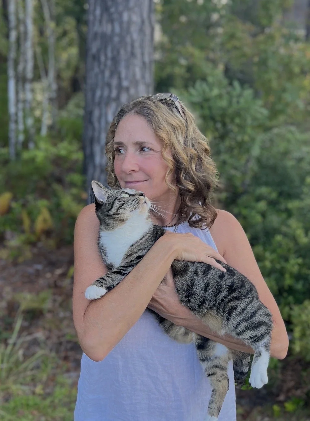 A woman holding a tabby cat outdoors in a green wooded area.