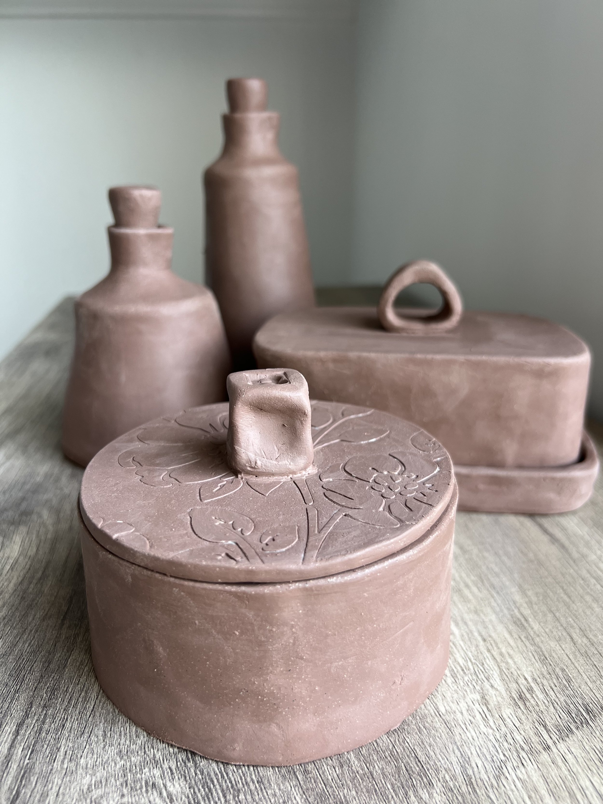 Multiple pinkish-brown ceramic vases and containers of various shapes and sizes on a wooden surface, against a light green wall.