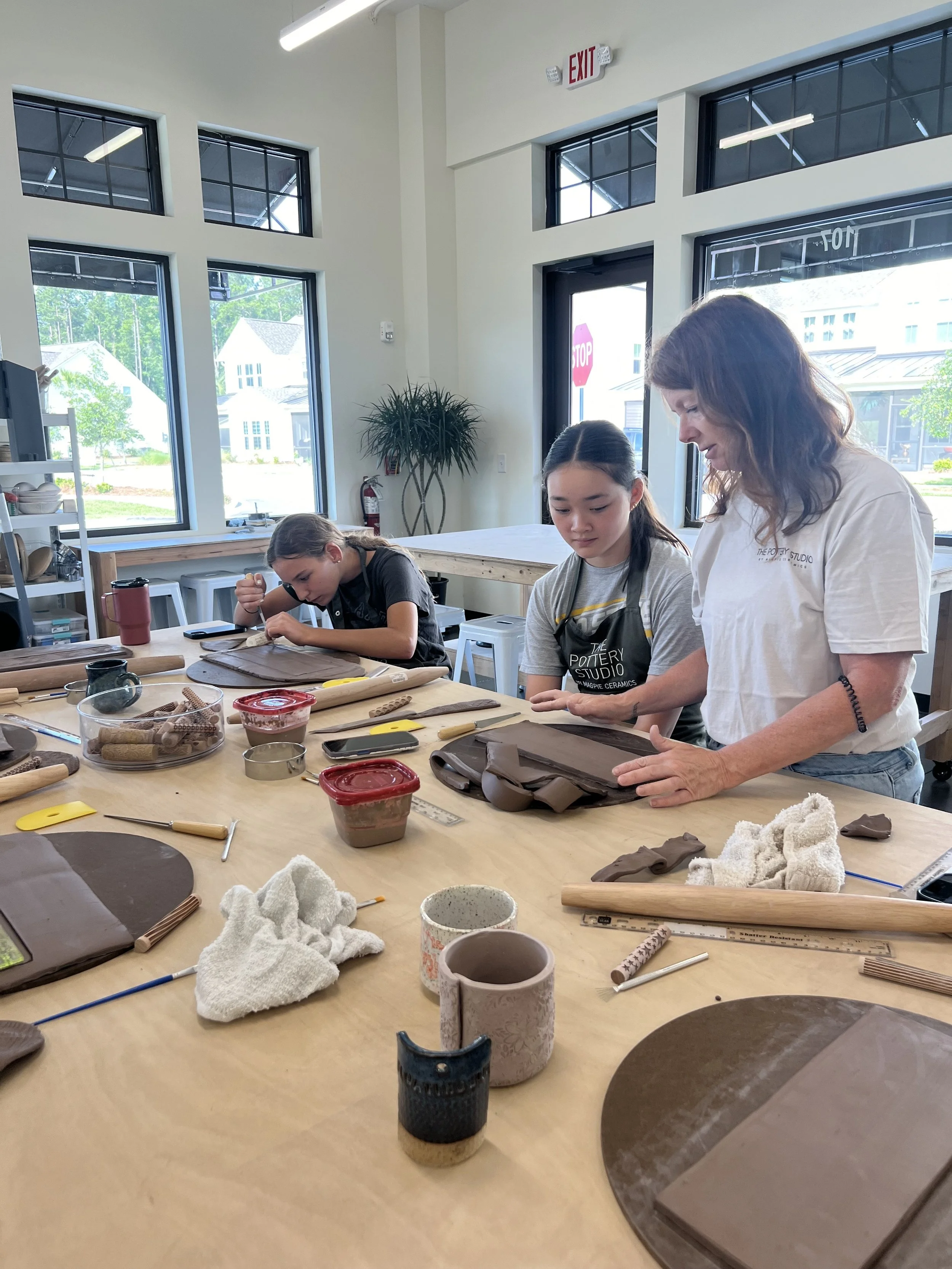 A pottery studio with three women working on their clay projects at a large table. The studio has bright natural light coming through large windows.