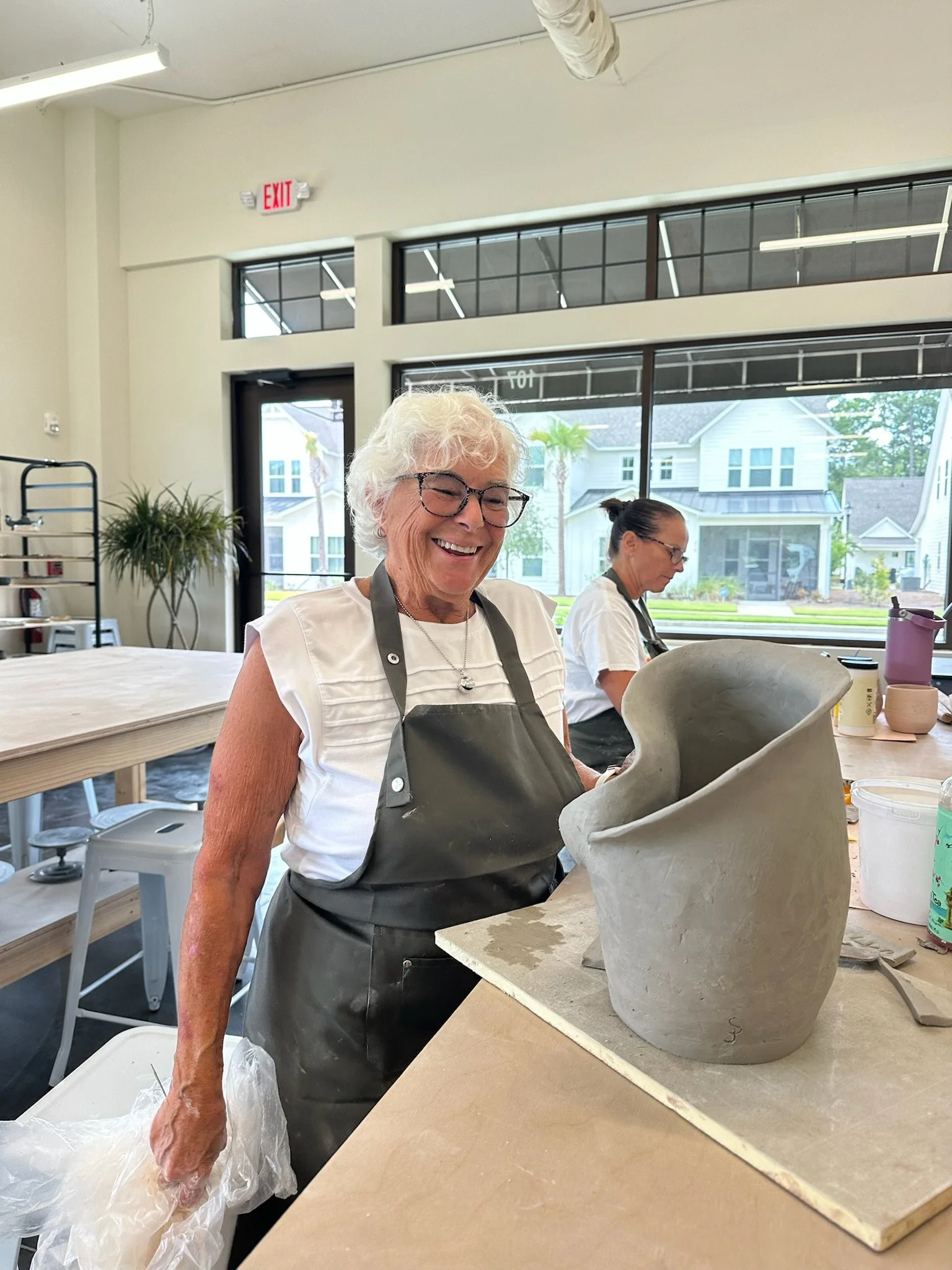 A smiling elderly woman with glasses, wearing a gray apron over a white shirt, working on a large ceramic piece in a well-lit studio with large windows and another woman in the background.