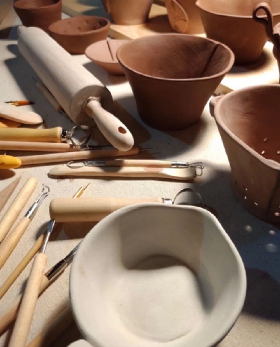Collection of pottery tools and several ceramic bowls and cups in various stages of completion on a worktable.