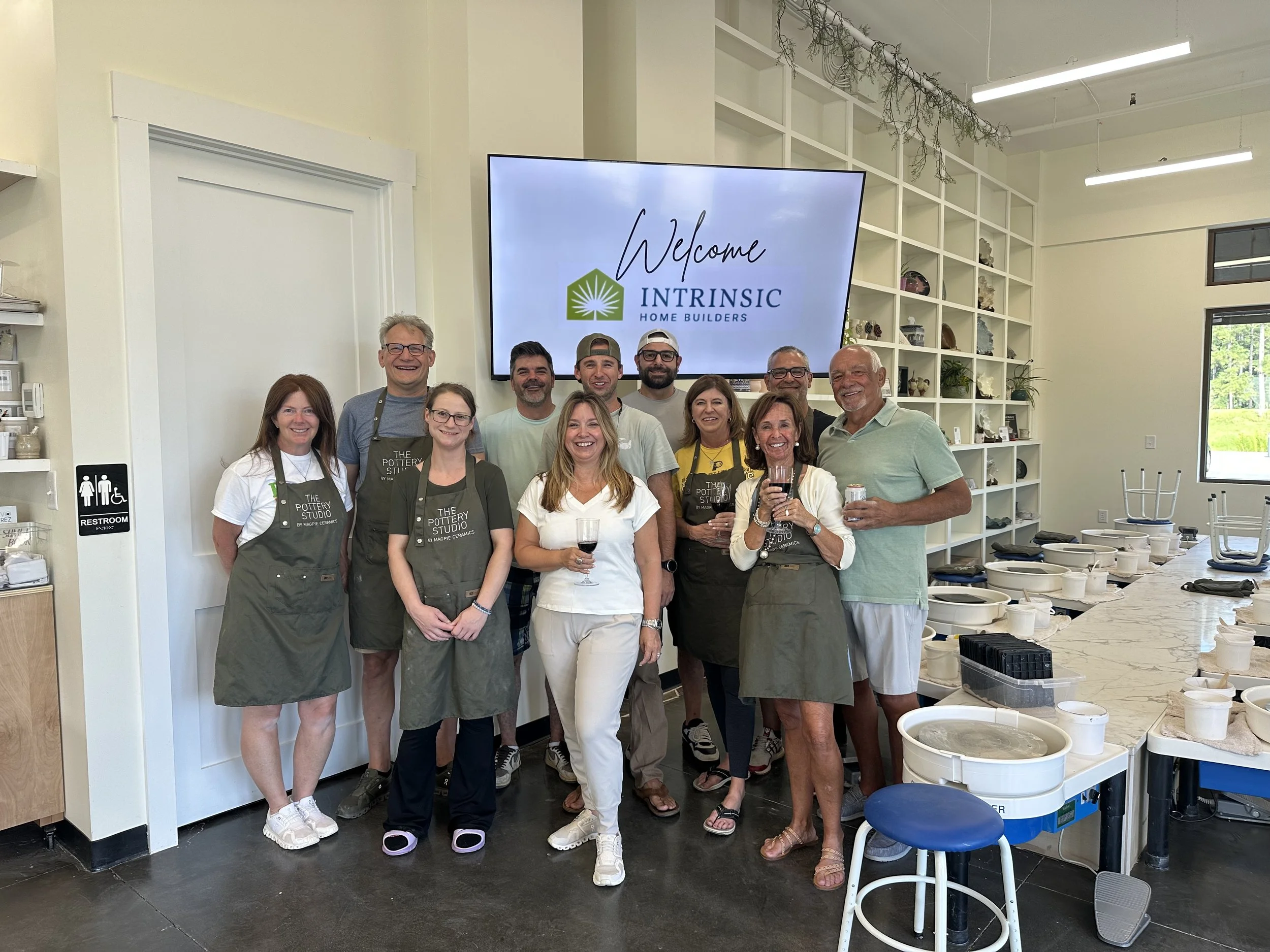 Group of people in a pottery studio, some wearing aprons, standing together and smiling at the camera, with pottery equipment and finished pieces on shelves behind them.