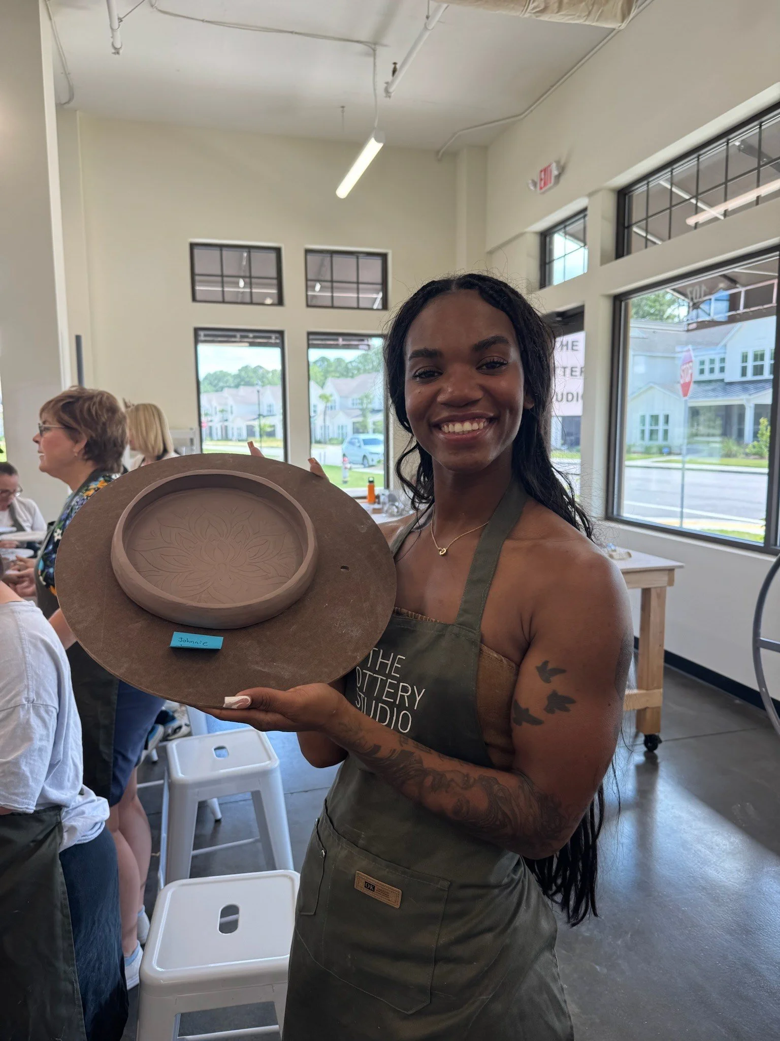 A woman holding a ceramic plate in a pottery studio, smiling at the camera. She is wearing an apron with the studio's name on it, has tattoos on her arm, and a necklace. Other people are present in the studio, and large windows outside show a residential neighborhood.