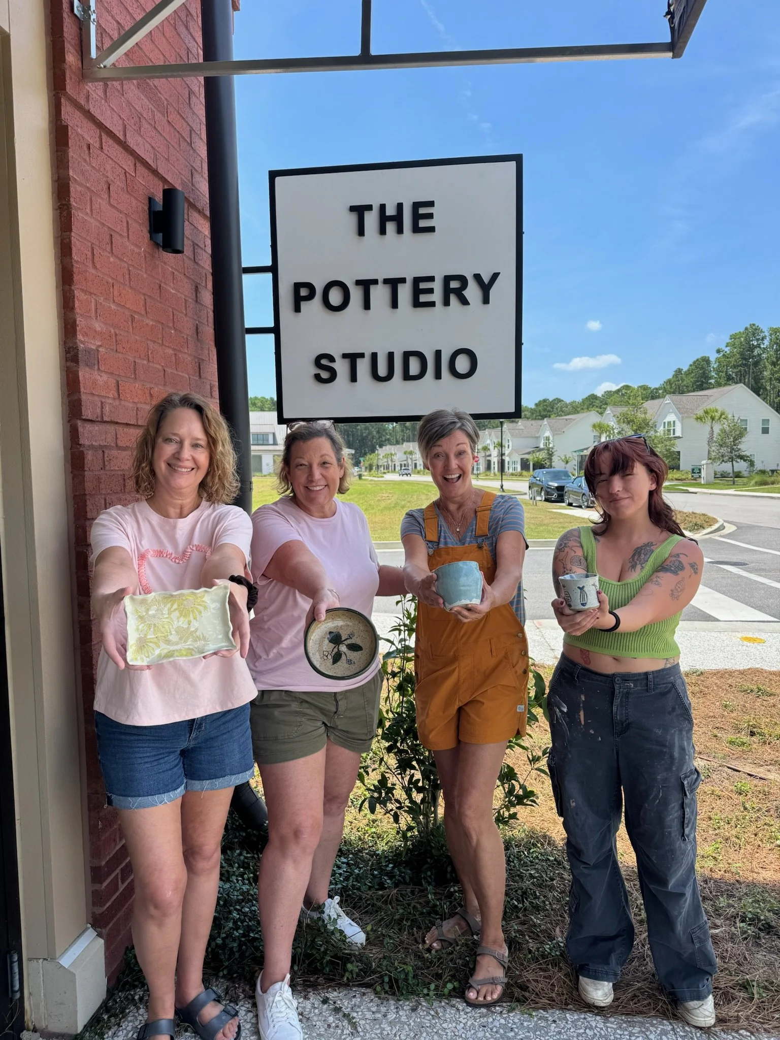 Four women standing outside in front of a sign that reads 'The Pottery Studio,' each holding a pottery piece they made.